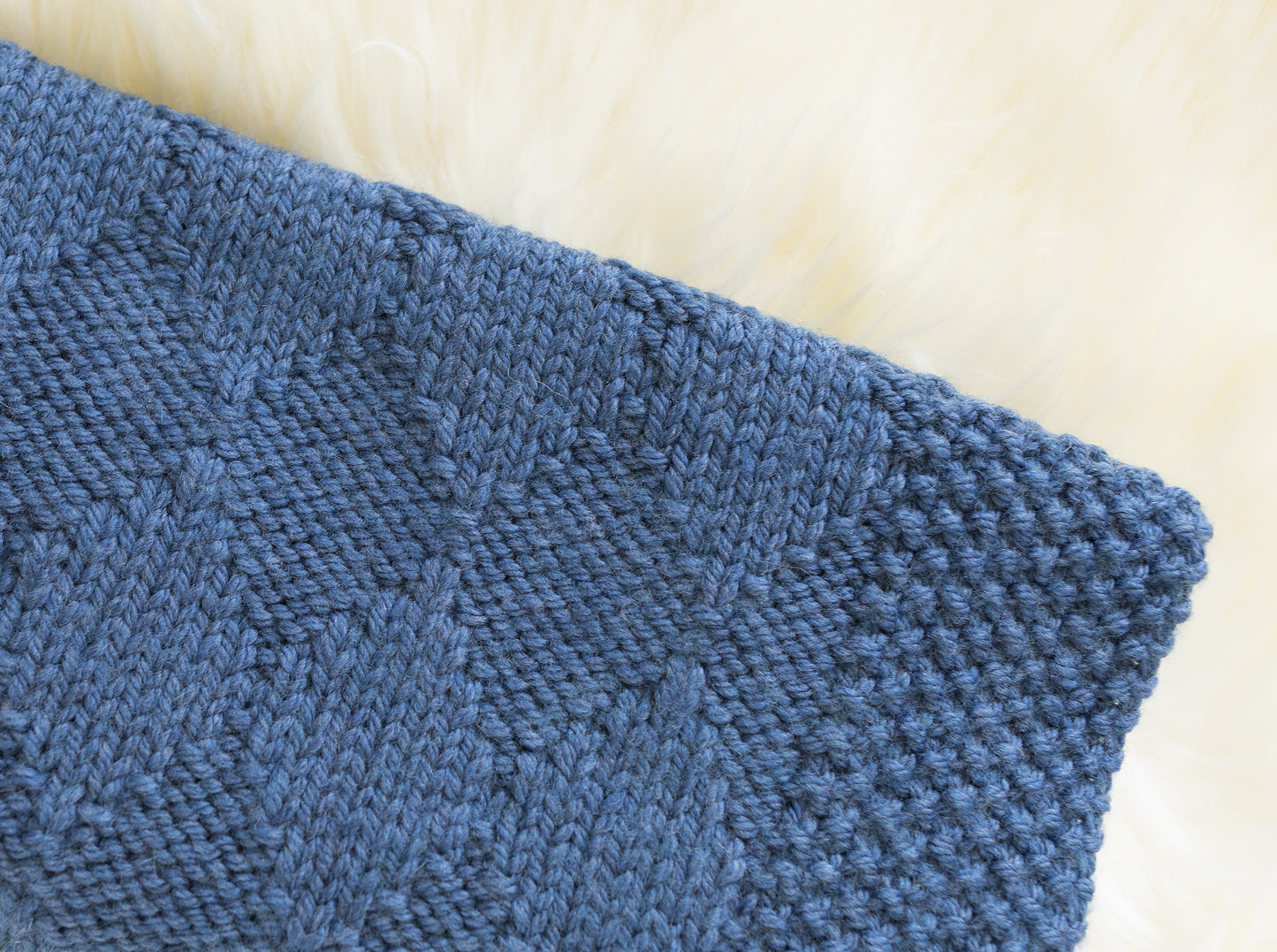A close-up of a textured blue knitted baby blanket with a diamond pattern, laid flat on a soft white fur background.