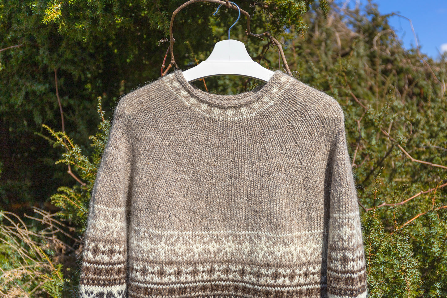 A hand-knit crewneck sweater in a heathered taupe-gray color with a white and dark brown Fair Isle pattern on the sleeves and hem, hanging on a white plastic hanger against a background of green bushes.