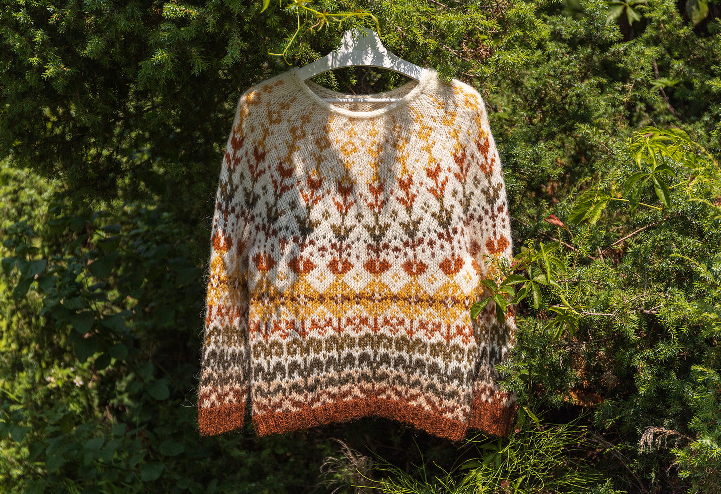 A long-sleeve knit sweater with a Fair Isle style pattern in cream, rust orange, mustard yellow, and dark gray, hanging on a clear plastic hanger against a background of green foliage.