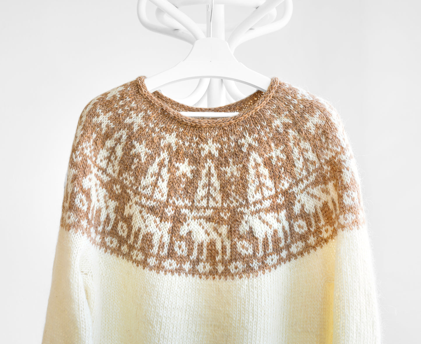 A cream-coloured knit jumper featuring a round yoke with a brown and white fair isle pattern depicting stylised mooses, trees, and stars.