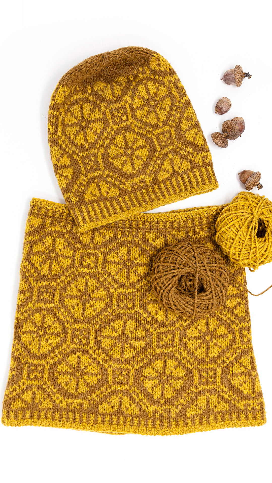Ratas Fair Isle hat and matching cowl with carriage wheel motif laid flat on a white background, both in a mustard yellow and dark brown patterned knit. Two balls of yarn and a few acorns are placed beside them as styling props.
