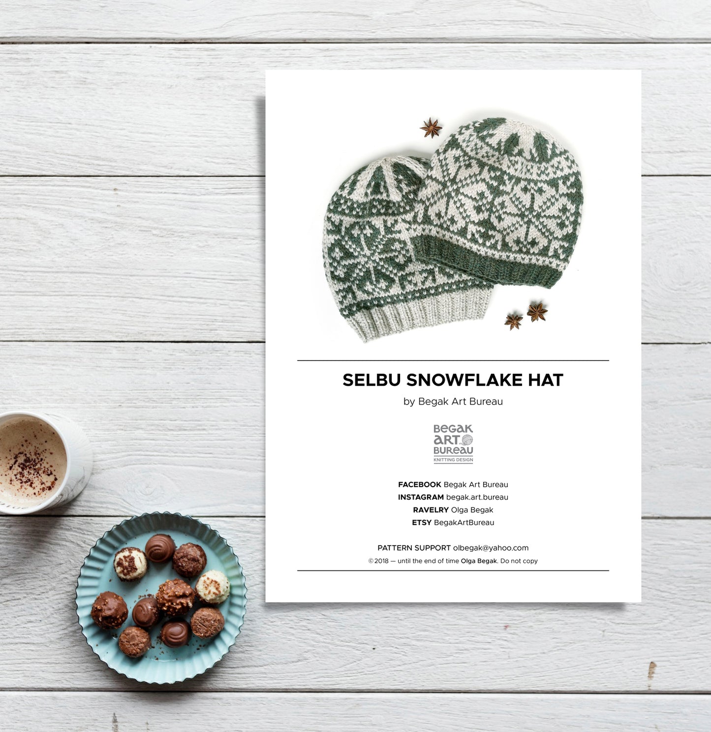 A flat-lay presentation of a knitting pattern card for the 'Selbu Snowflake Hat' by Begak Art Bureau. The white card features a green and white hand-knit snowflake hat illustration, surrounded by decorative props including a cup of coffee, a small plate of chocolate truffles, and star anise, all arranged on a light gray wood plank background.