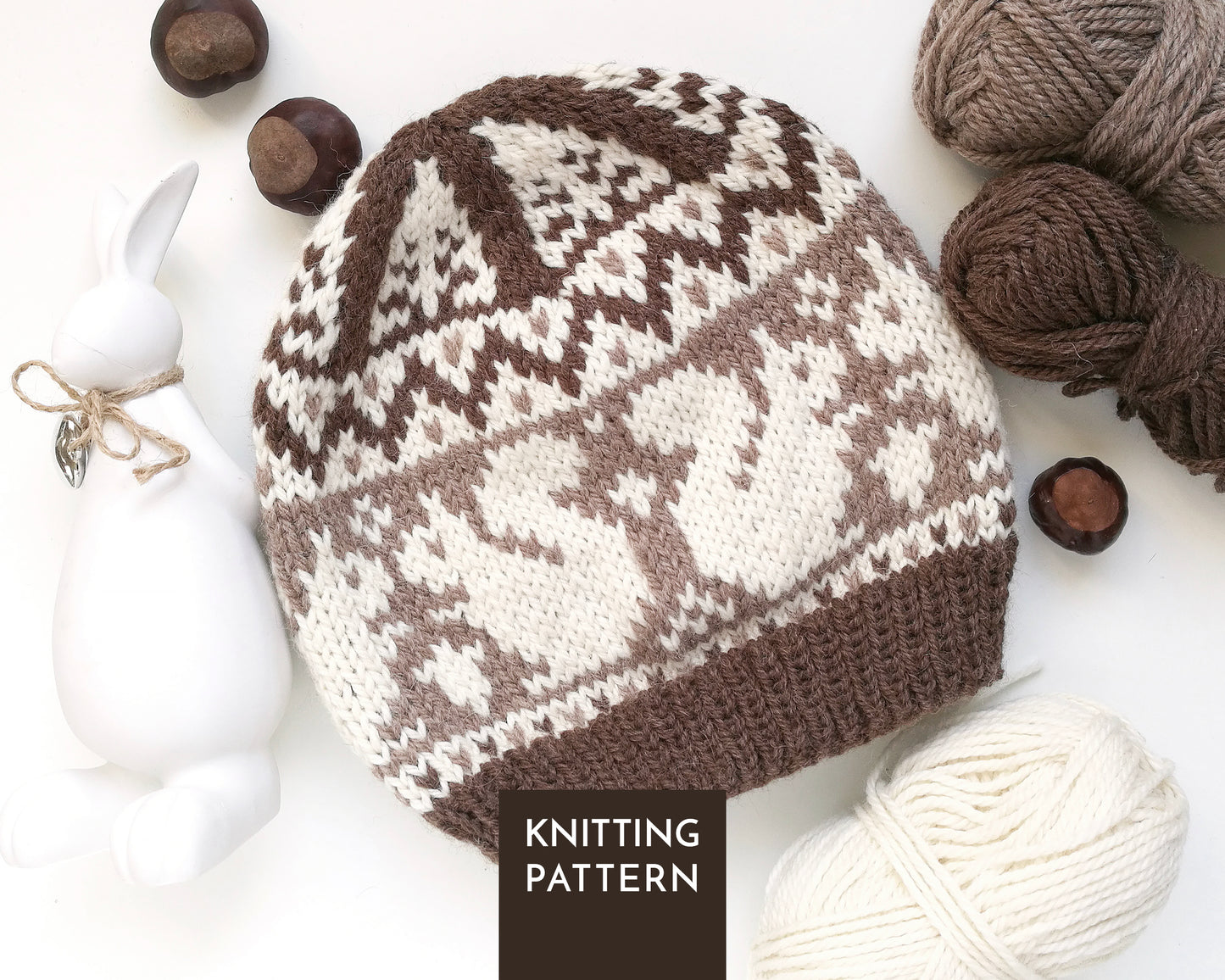 A knitted beanie hat with a brown and cream Squirrel Fair Isle pattern, styled in a flat lay with yarn balls, chestnuts, a white ceramic rabbit figurine.