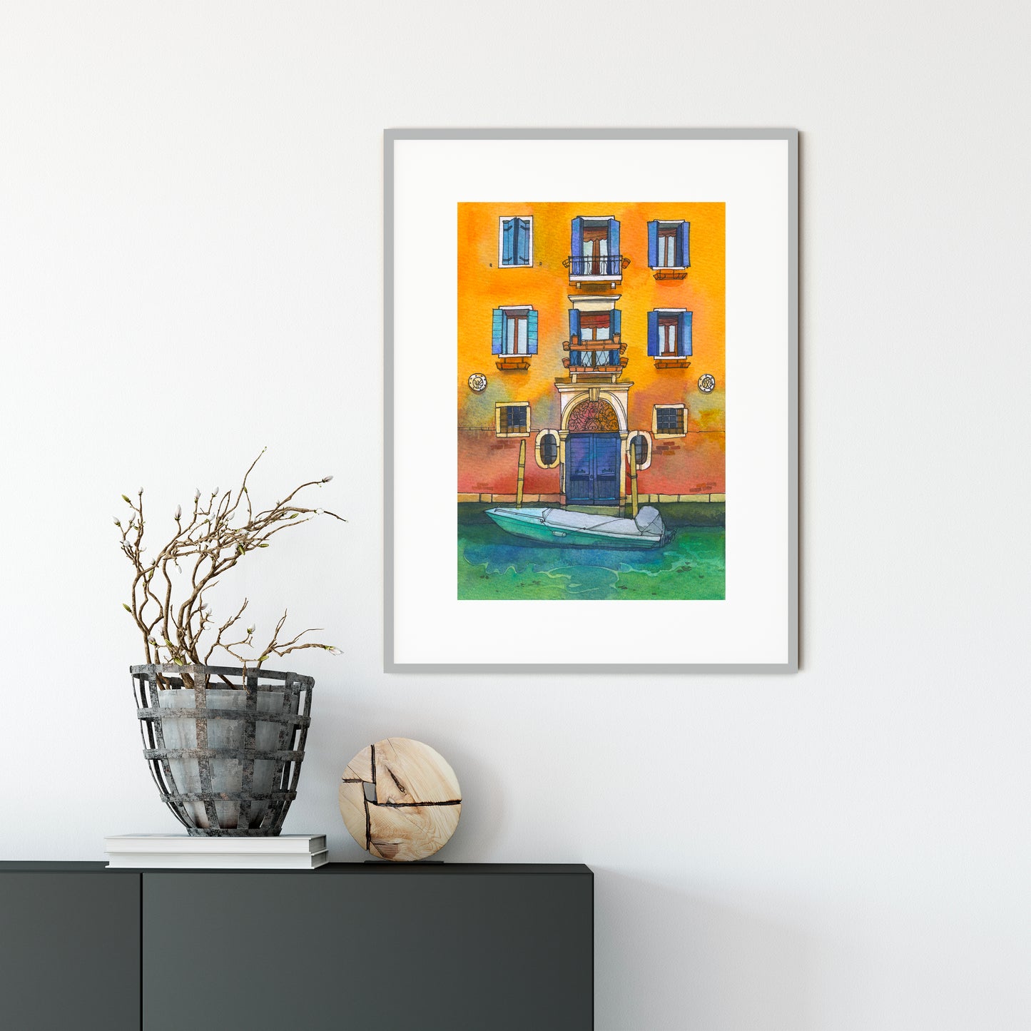 Framed watercolor artwork of a Venetian canal scene with a yellow building and blue door, placed on a white wall above a dark surface.