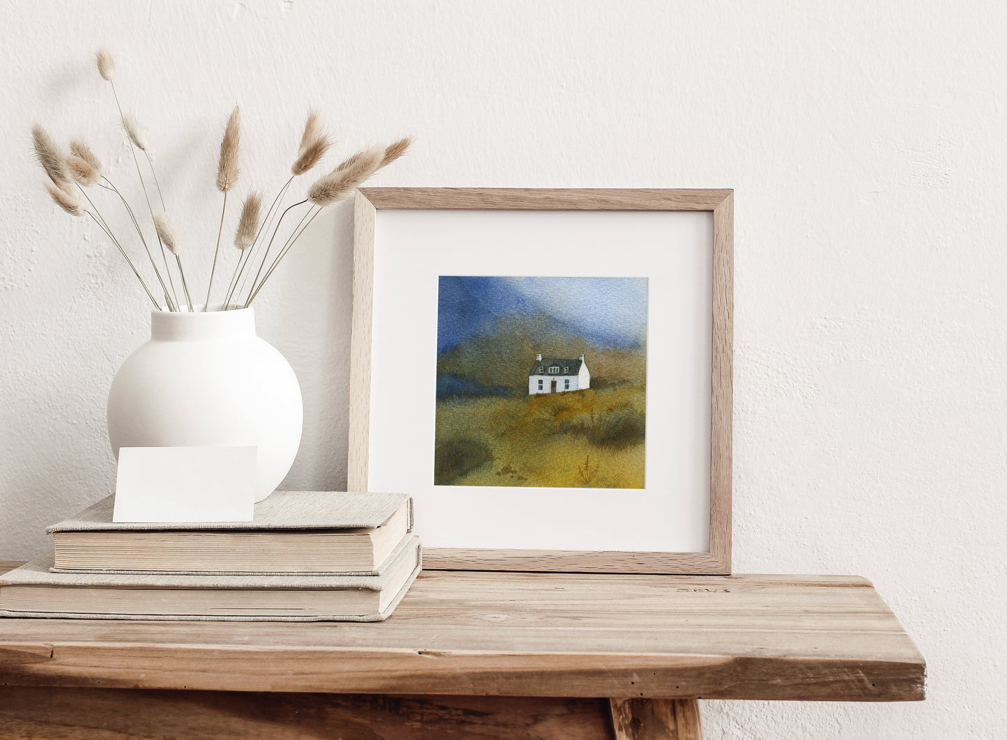 Framed watercolor painting featuring a small white Allt Dearg cottage sits below the Skye Ridge on the Isle of Skye, Scottish Highlands, displayed on a wooden surface with a vase and books