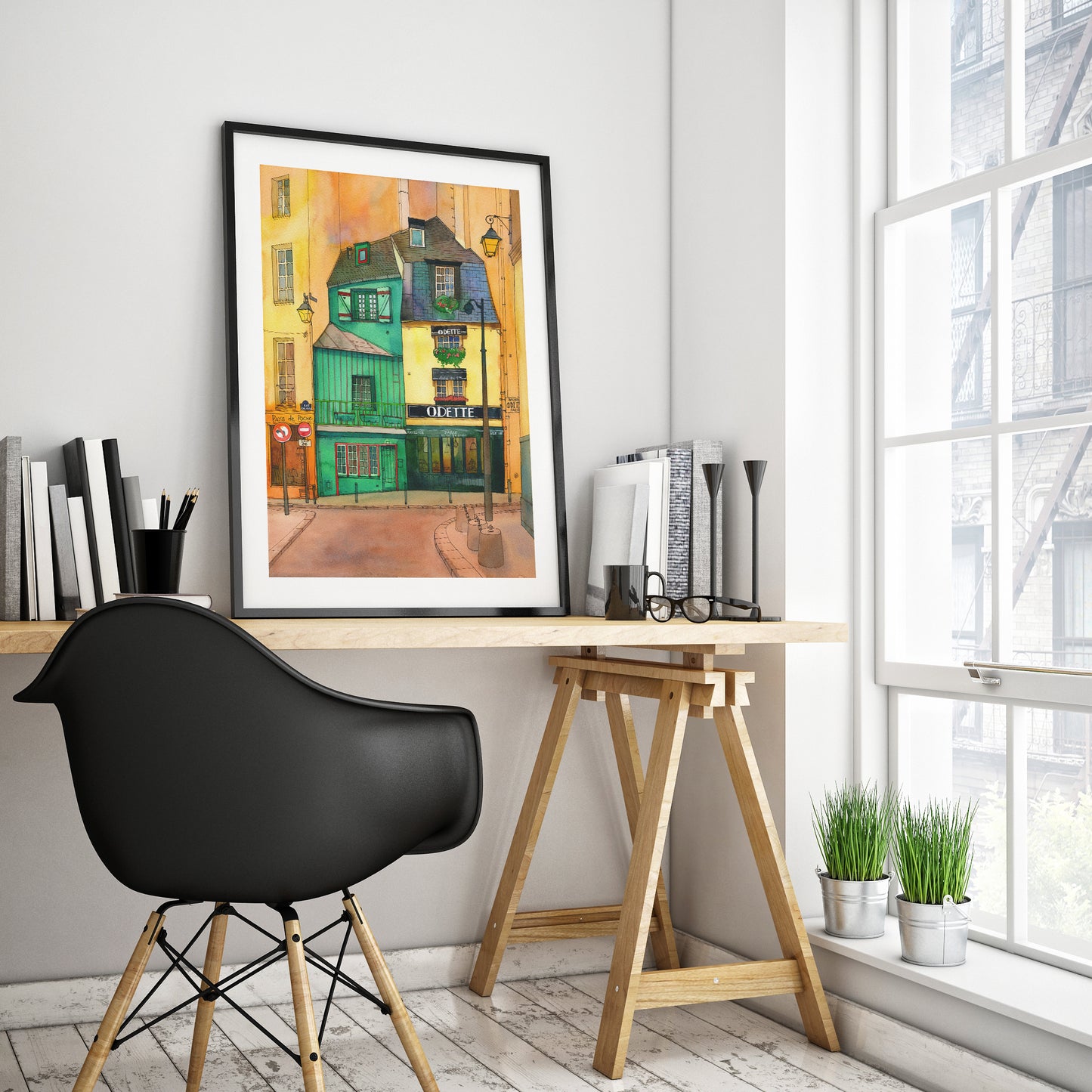 A framed Café Odette in Paris watercolor and ink art print, displayed on a wooden desk in a modern home office setting next to a window.
