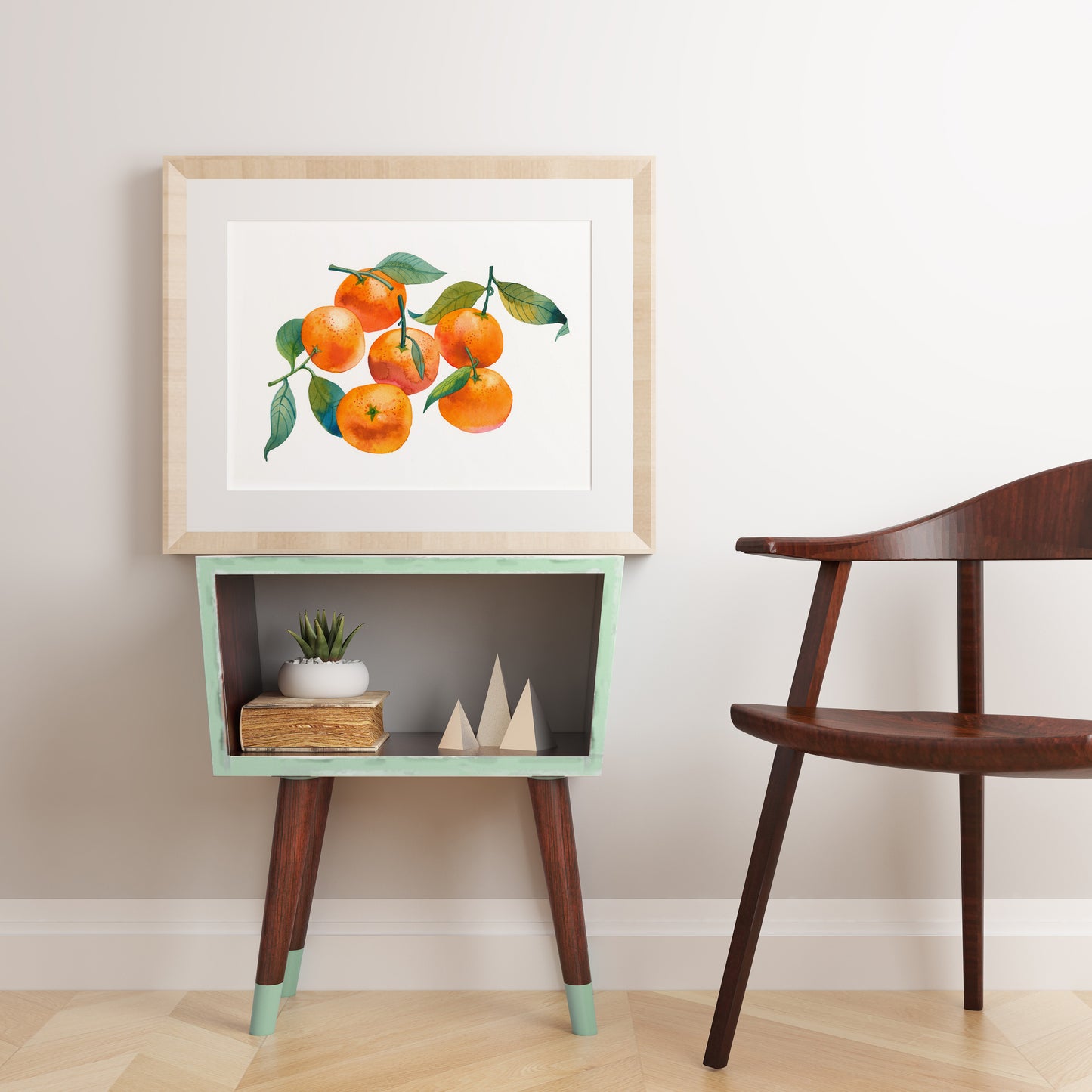 A framed watercolor art print of orange mandarins with green leaves, styled in a light wood frame and leaning on a mint green and dark wood side table next to a wooden chair.