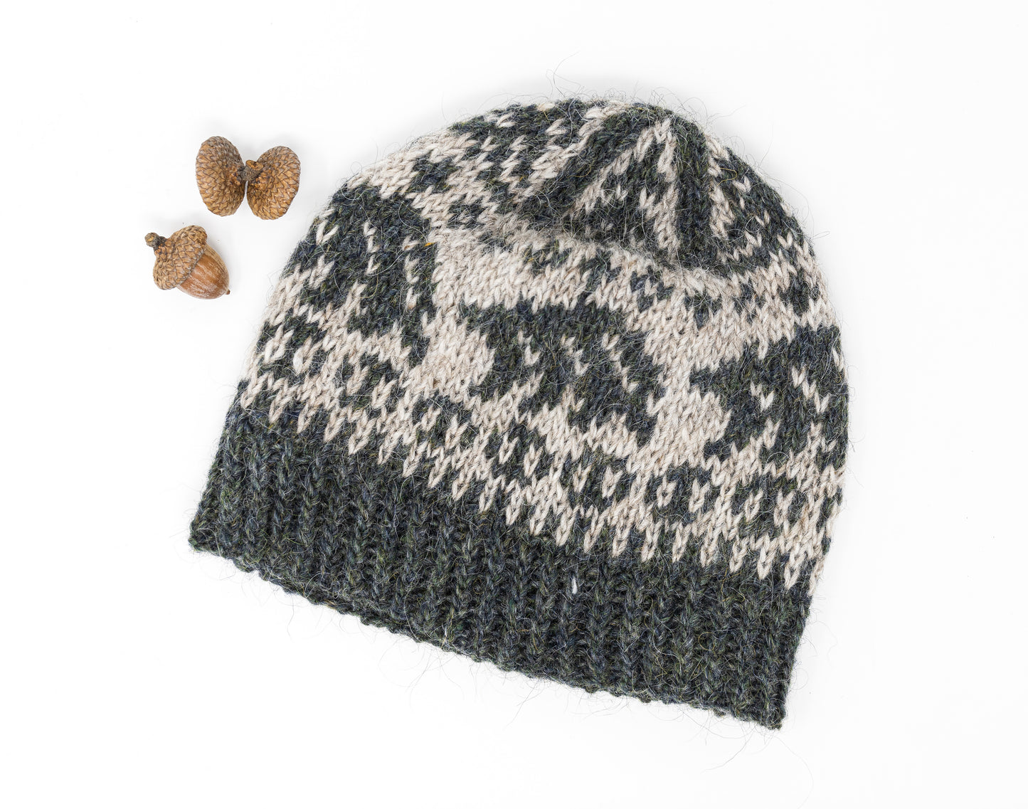 A hand-knit beanie hat with a dark gray-green and light beige Fair Isle style pattern, featuring bears, displayed on a white background next to small acorns.