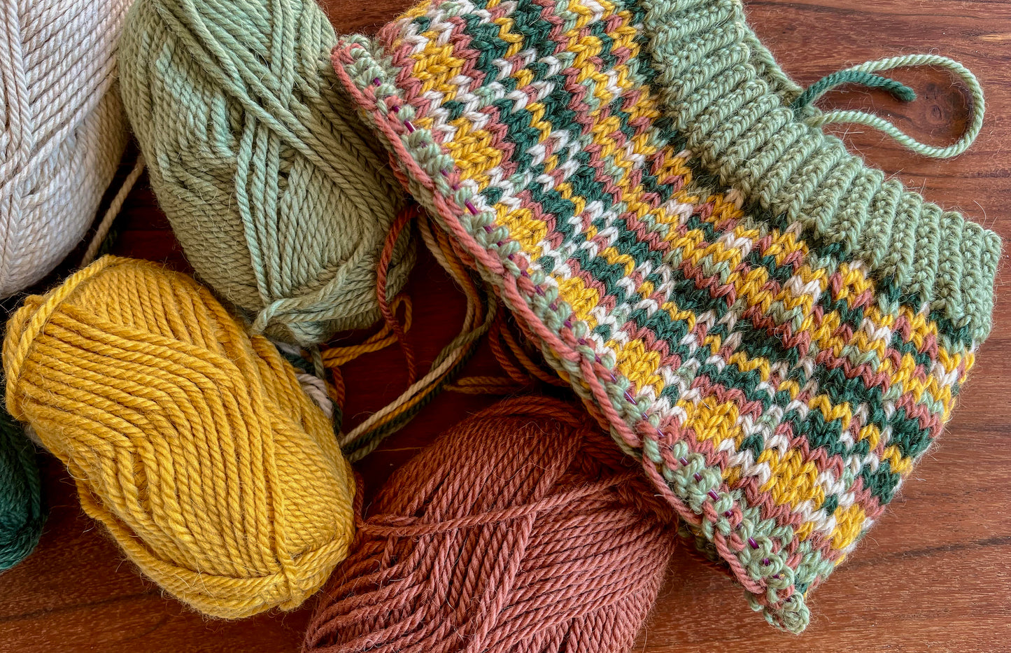 A hand-knitted hat with a Fair Isle pattern in dark green, sage green, yellow, pink, and beige in progress, displayed on a wooden surface next to several skeins of yarn.