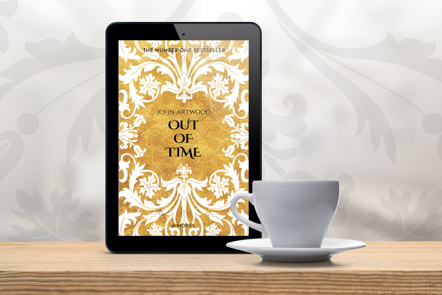 A tablet with a book cover featuring gold and white baroque ornament, accompanied with a white cup on a wooden table.