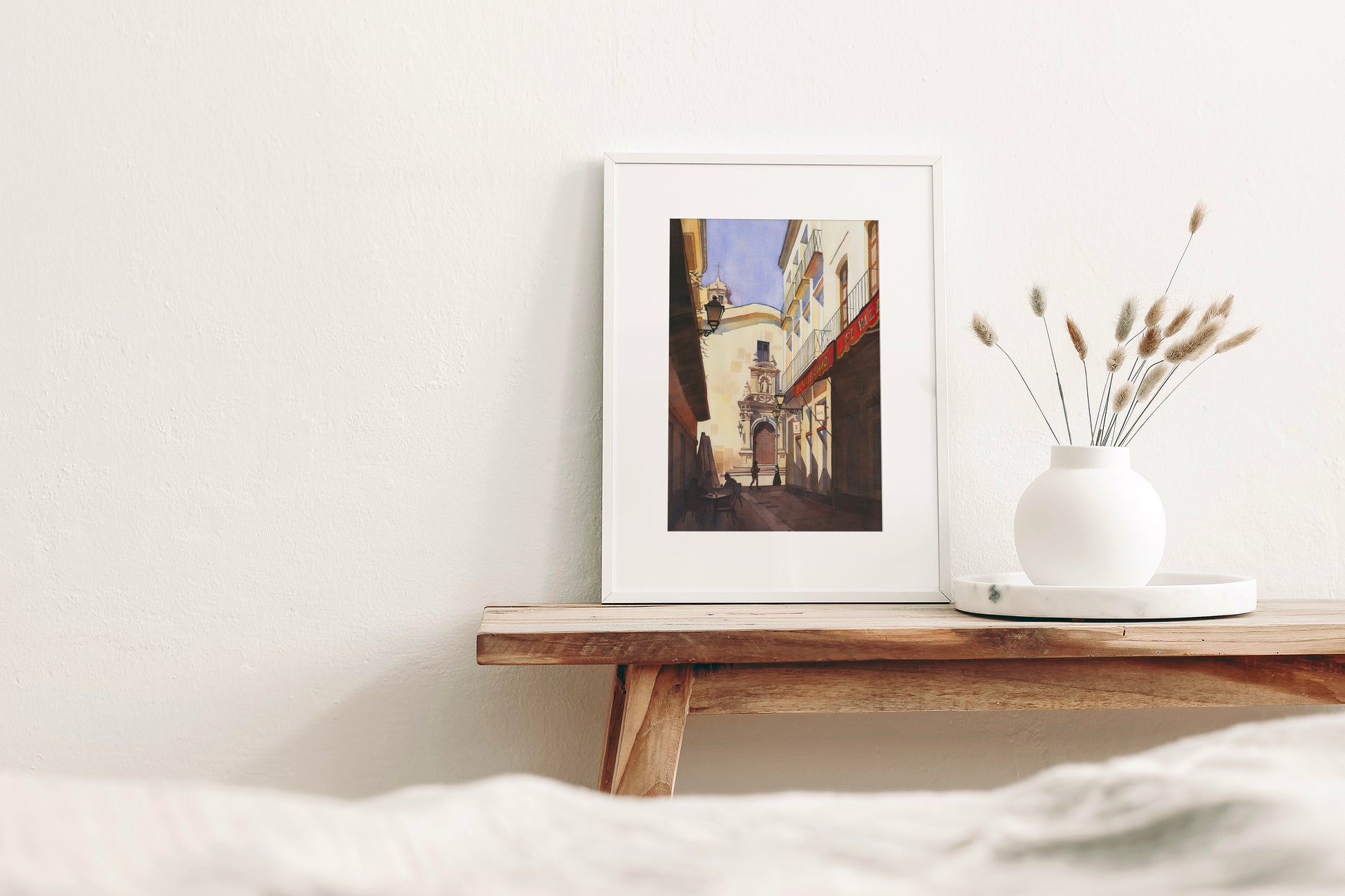 framed watercolor artwork of a street in Almeria Old town, Spain, on a wooden shelf with a vase of dried plants against a white wall.