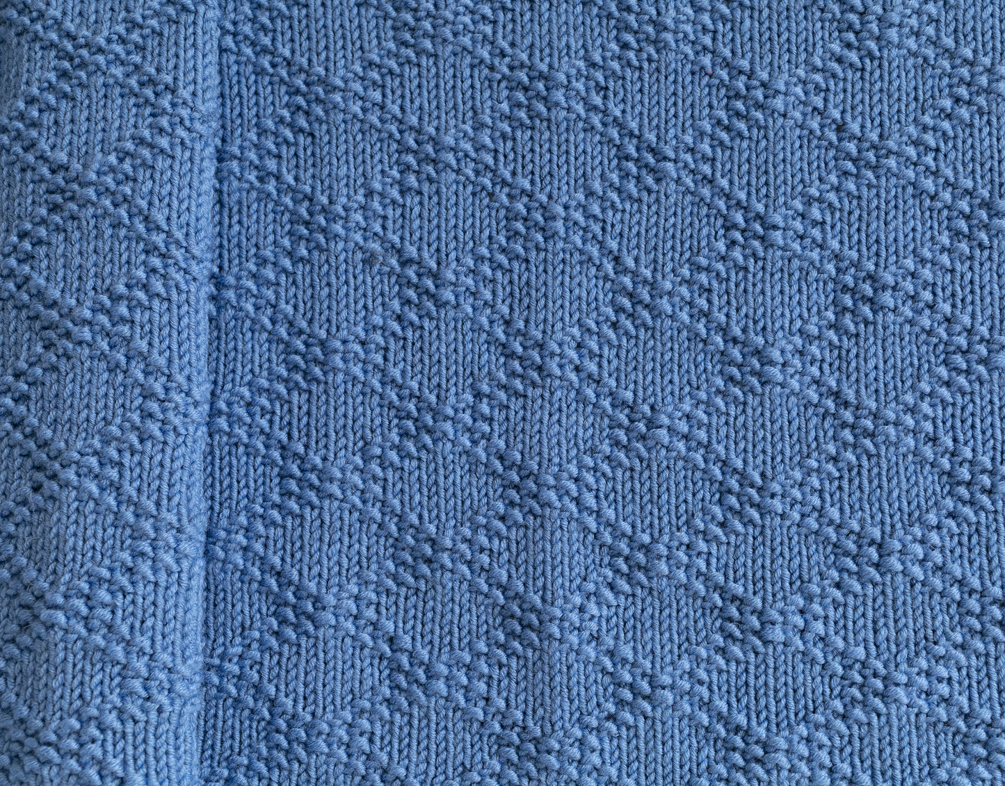 A close-up of a textured knit blanket in blue color, showing a repeating Charles Brocade pattern.