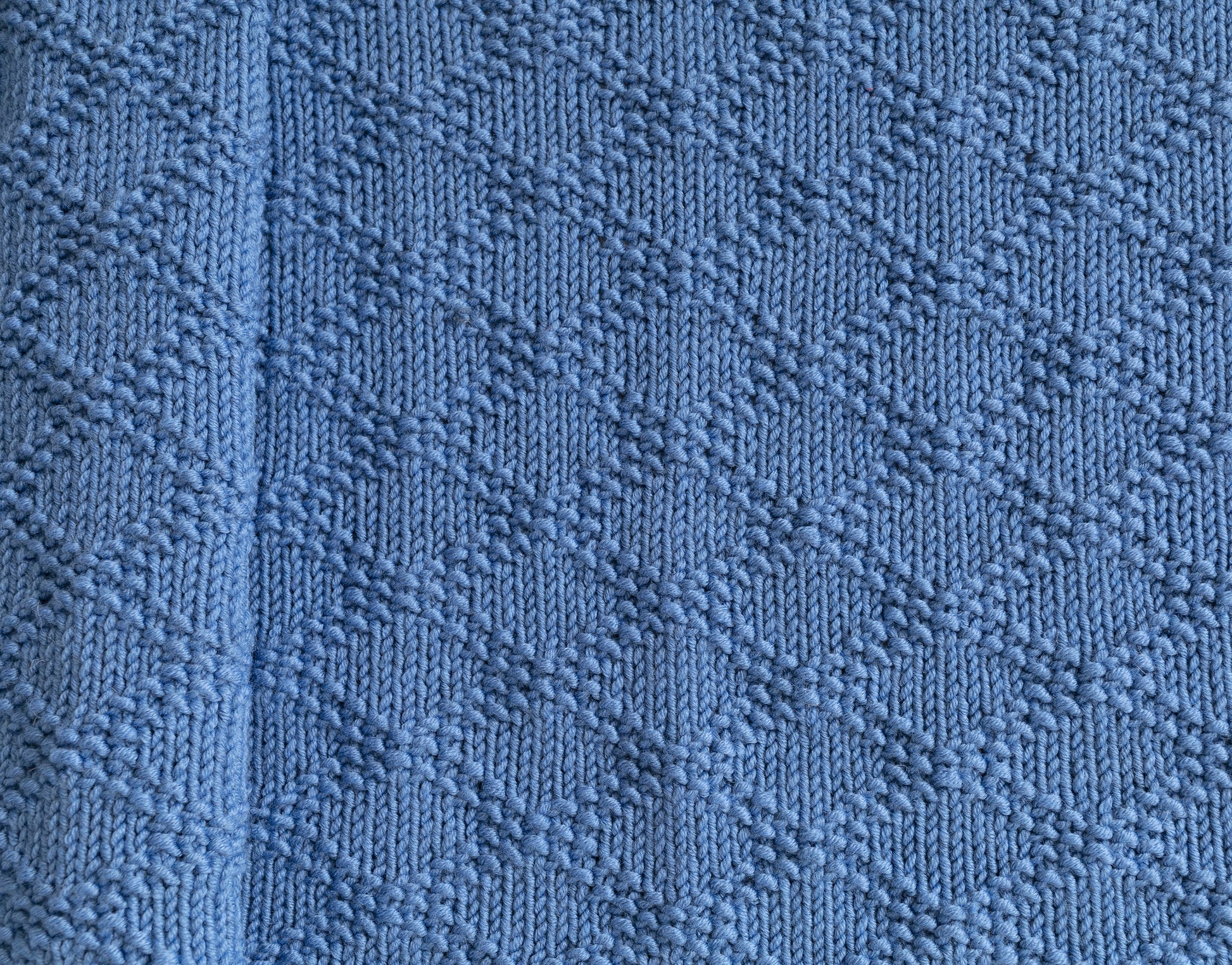 A close-up of a textured knit blanket in blue color, showing a repeating Charles Brocade pattern.