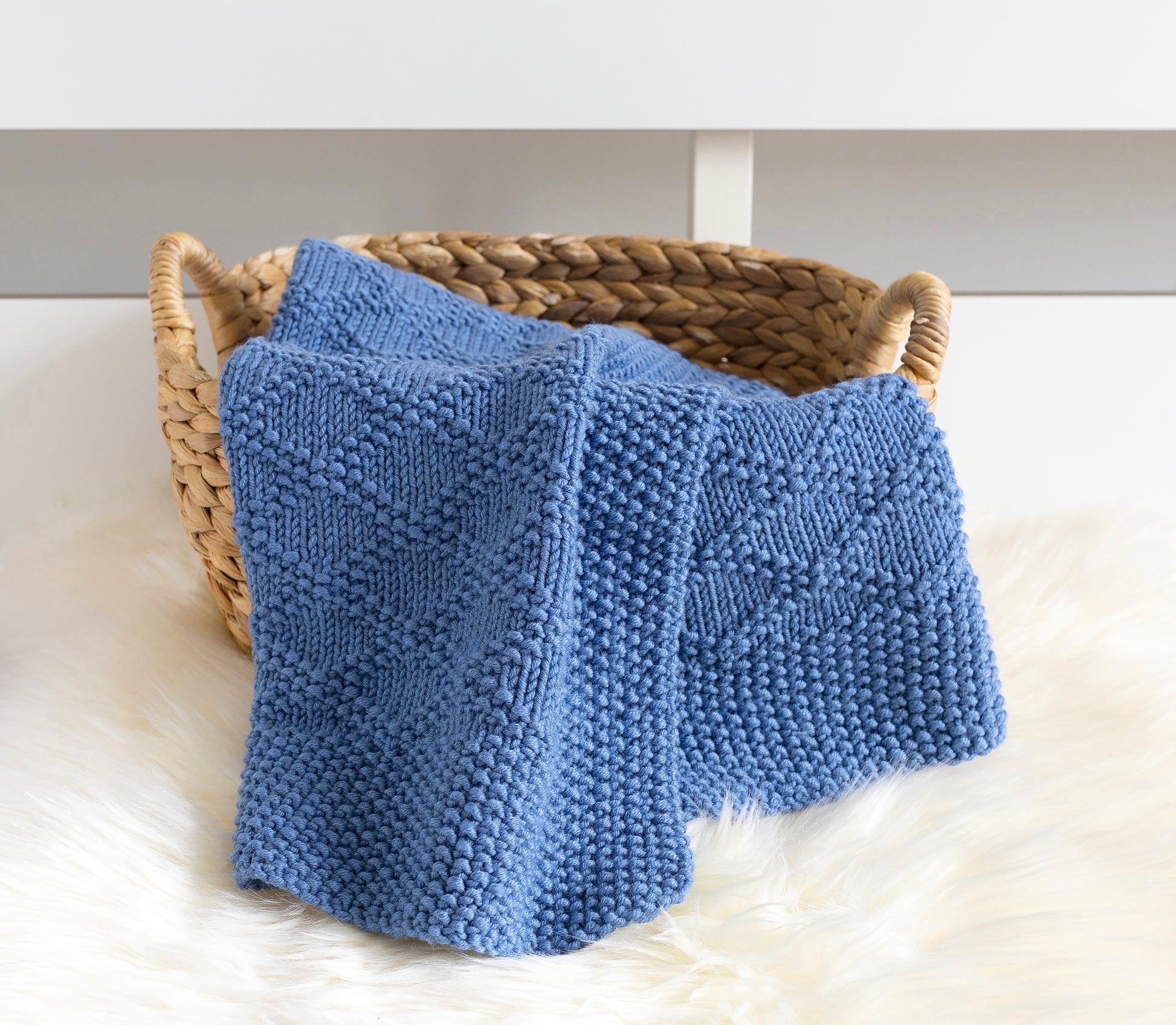 A blue knitted blanket with textured Charles Brocade patterns draped over a woven straw basket on a white fur surface.