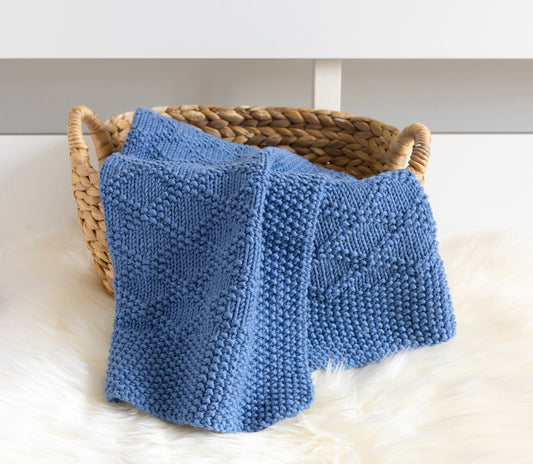 A blue knitted blanket with textured Charles Brocade patterns draped over a woven straw basket on a white fur surface.