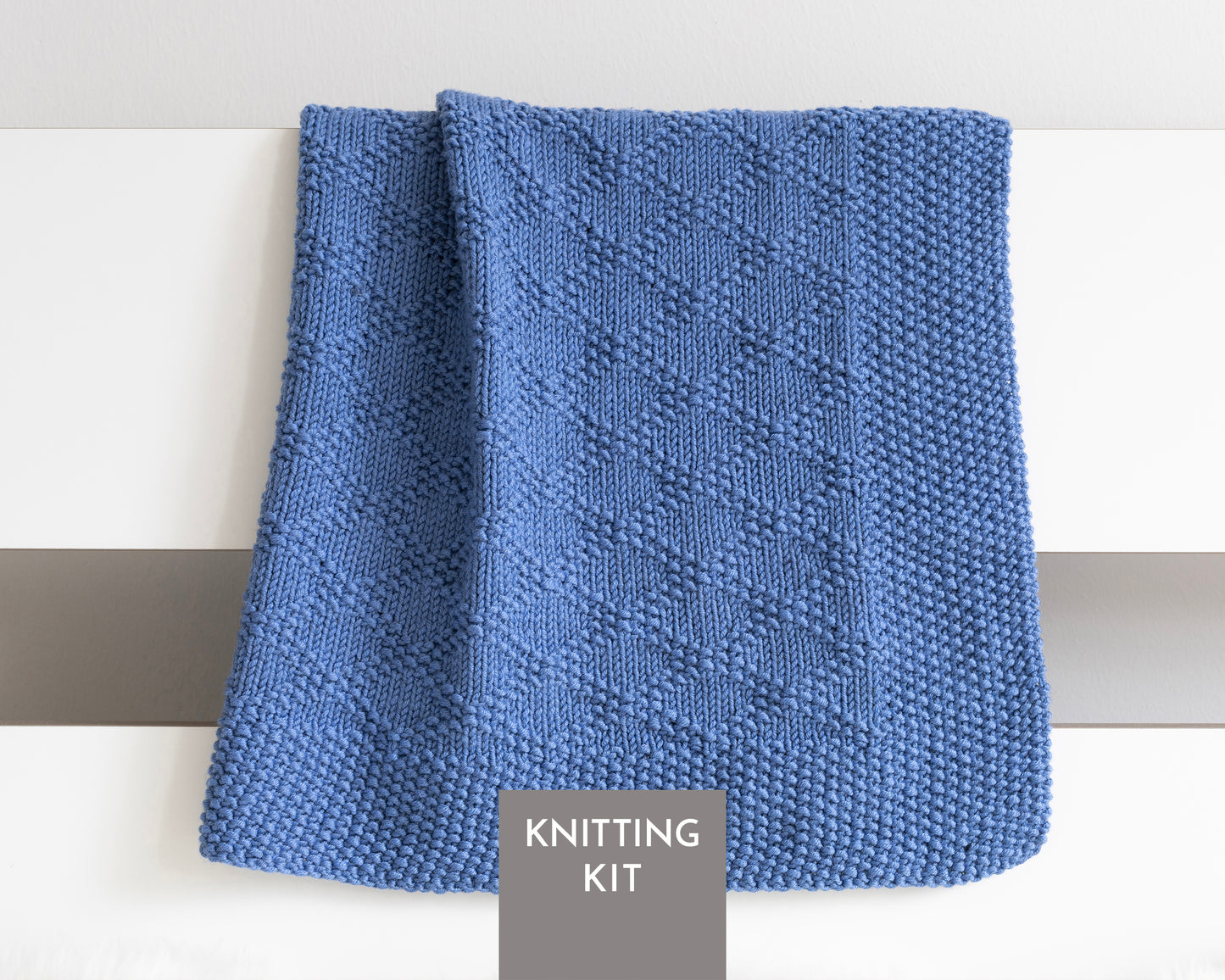 A blue knitted textured blanket with Charles Brocade pattern, draped over a white surface with a gray label reading 'KNITTING KIT' at the bottom.