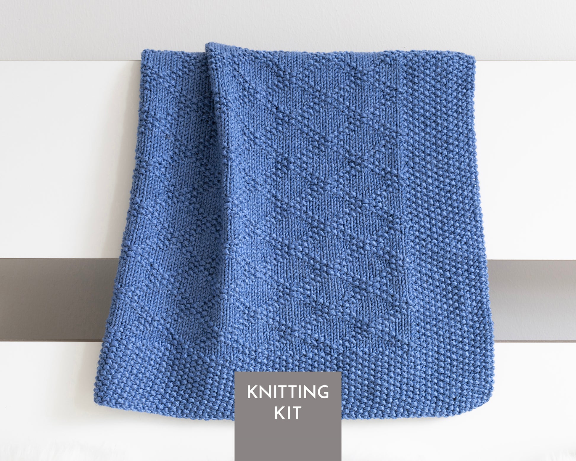 A blue knitted textured blanket with Charles Brocade pattern, draped over a white surface with a gray label reading 'KNITTING KIT' at the bottom.