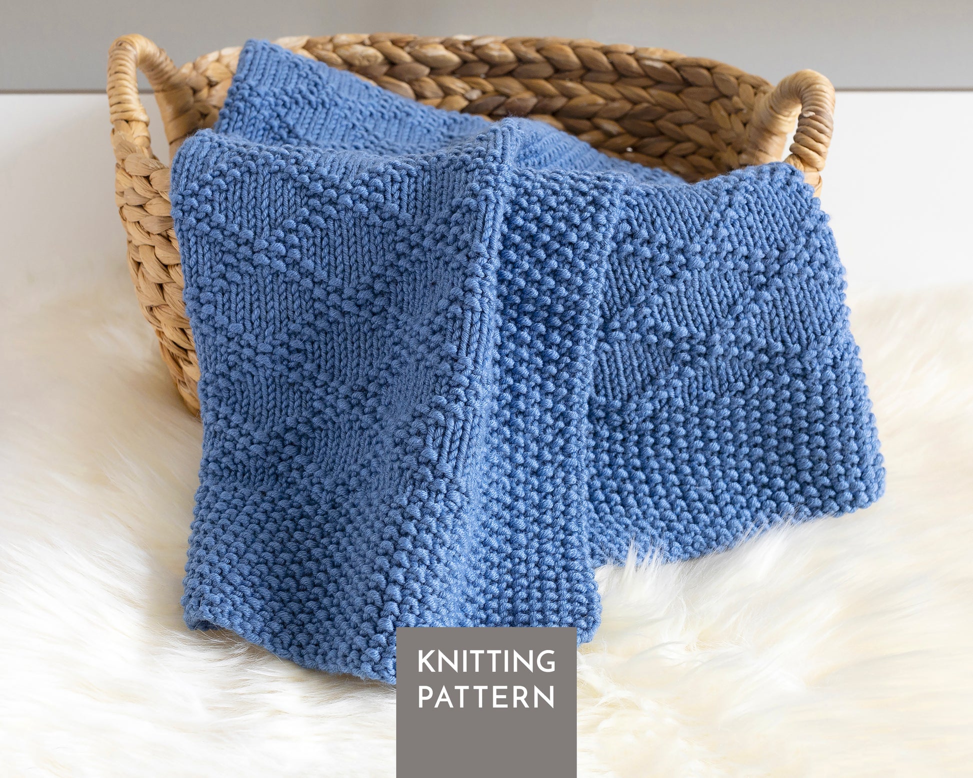 A blue knitted blanket with a textured Charles Brocade pattern draped over a woven wicker basket on a white faux fur surface. A gray label at the bottom reads 'KNITTING PATTERN'.
