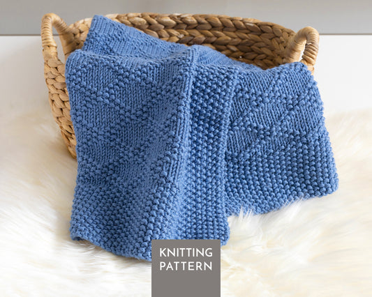A blue knitted blanket with a textured Charles Brocade pattern draped over a woven wicker basket on a white faux fur surface. A gray label at the bottom reads 'KNITTING PATTERN'.