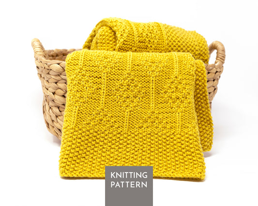 A knitted mustard yellow blanket displayed in a woven straw basket on a white background, with a gray label reading 'KNITTING PATTERN' at the bottom.