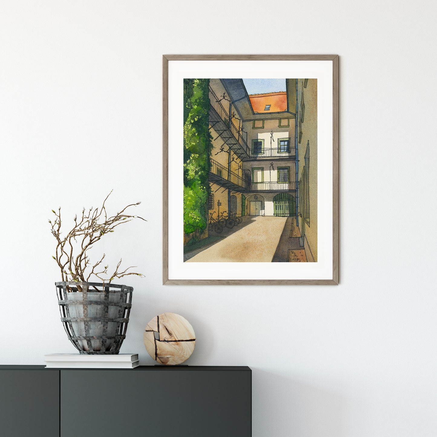 Framed watercolor and ink artwork of a Graz city street scene on a white wall with decorative items below.
