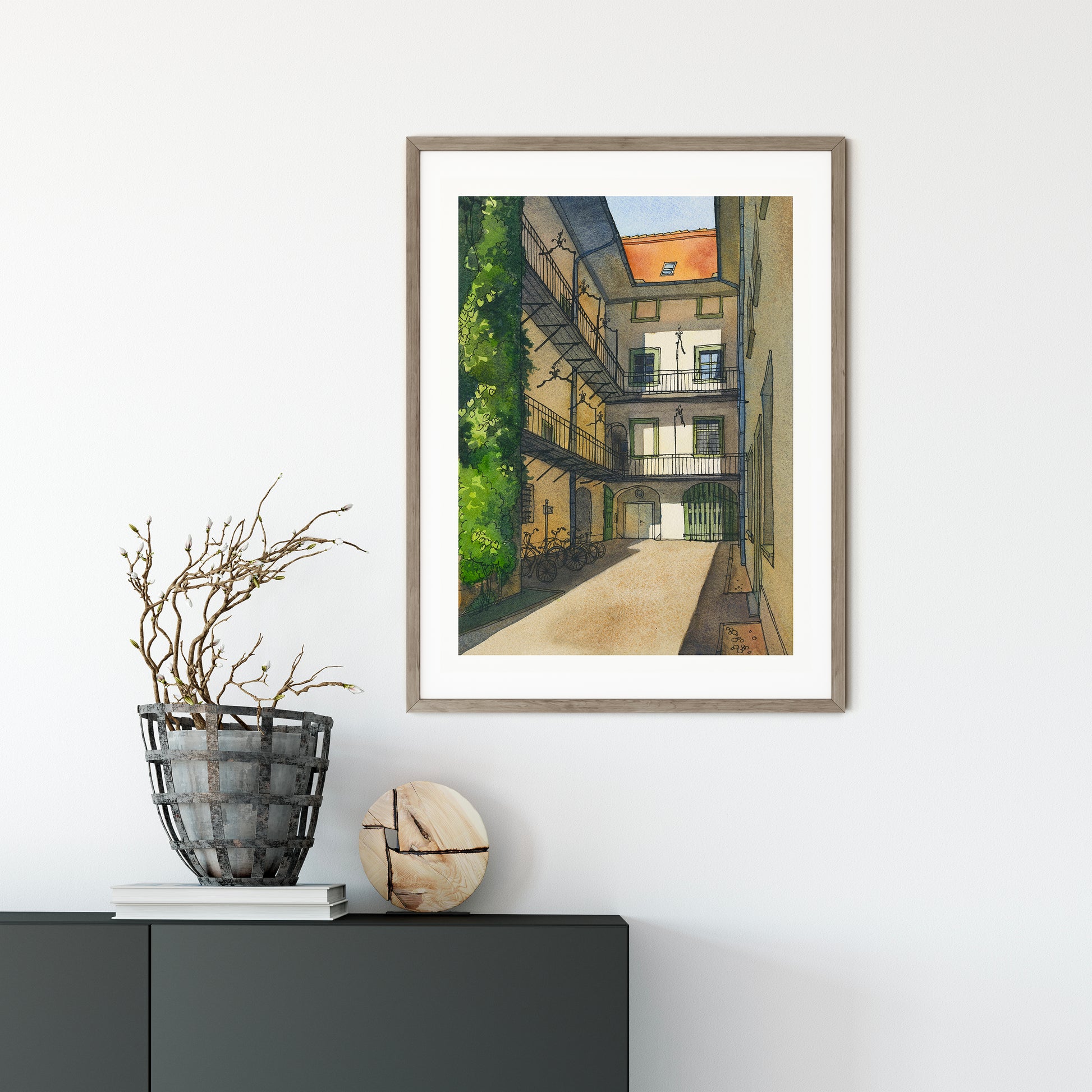 Framed watercolor and ink artwork of a Graz city street scene on a white wall with decorative items below.