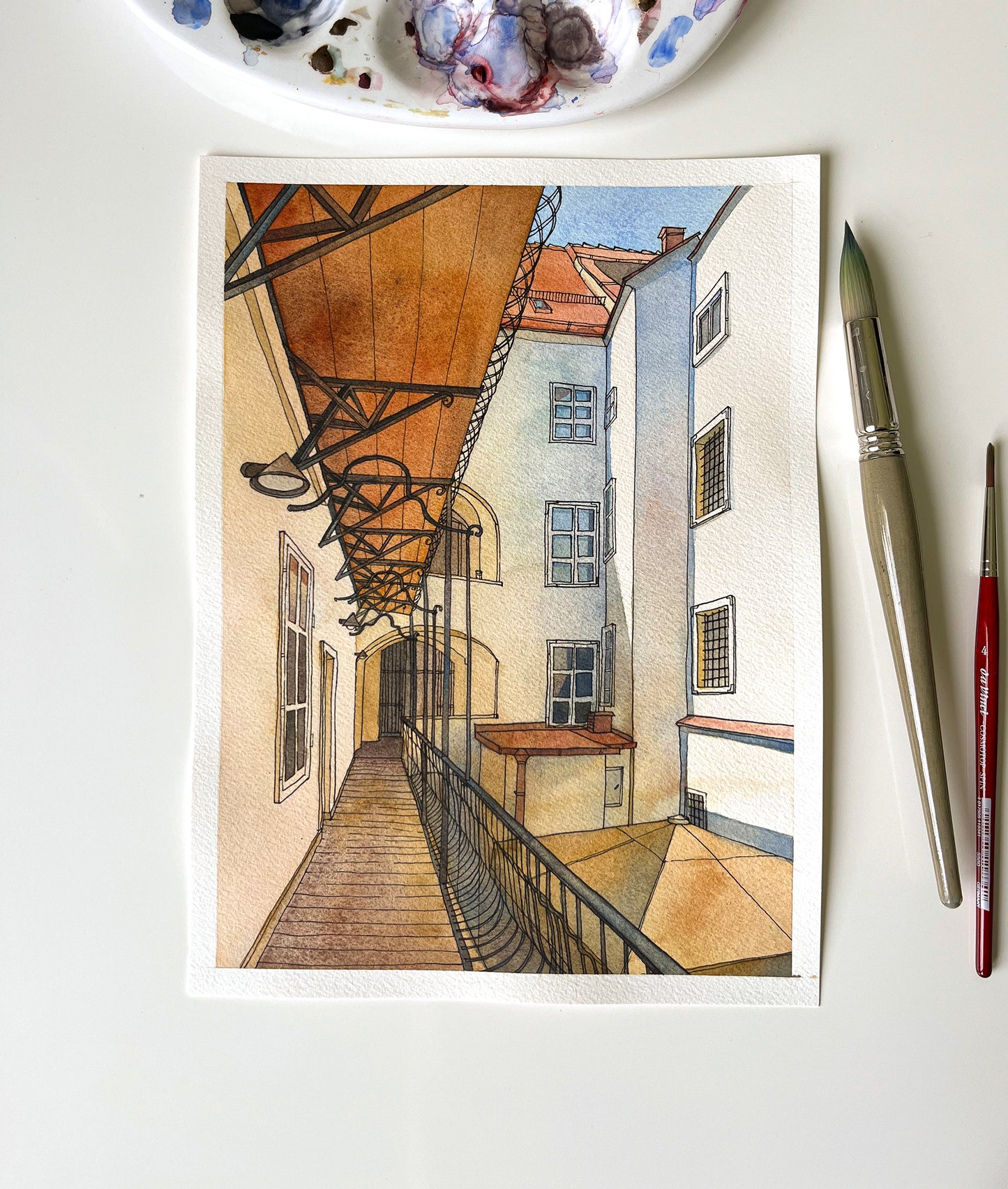 original watercolor and ink artwork of a historic courtyard in Graz on a white surface, accompanied by art supplies.