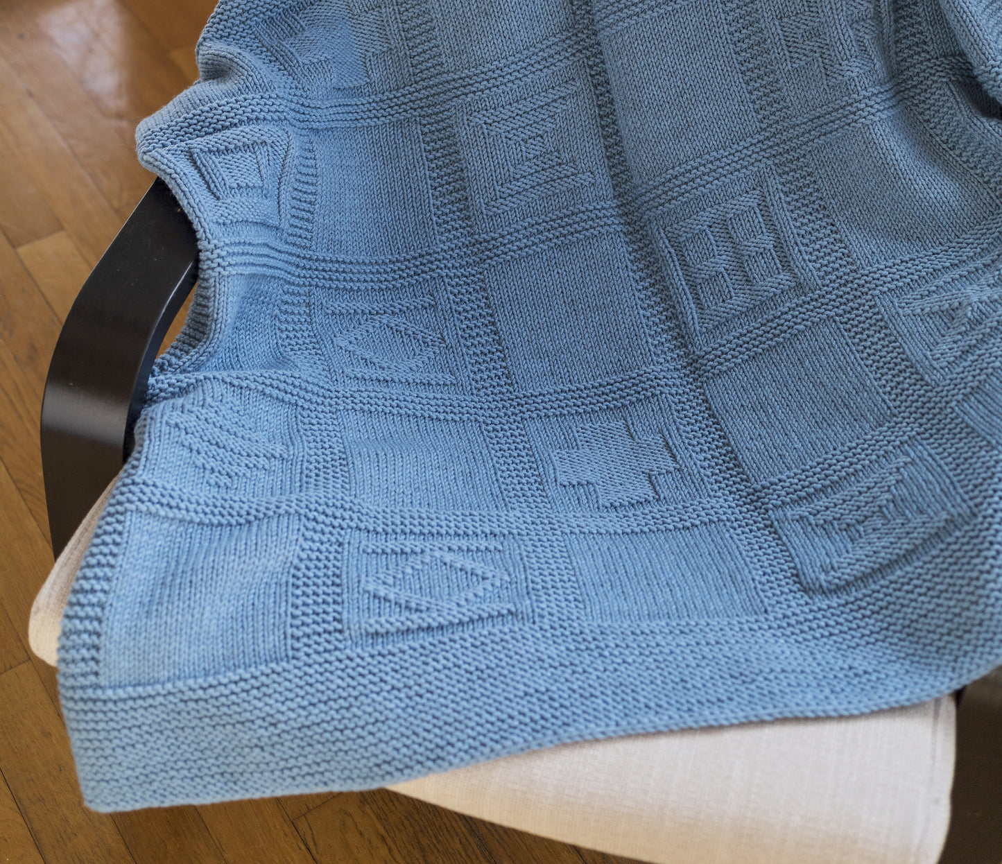 A blue merino wool hand-knitted baby blanket in geometric pattern draped on a armchair, Leland design