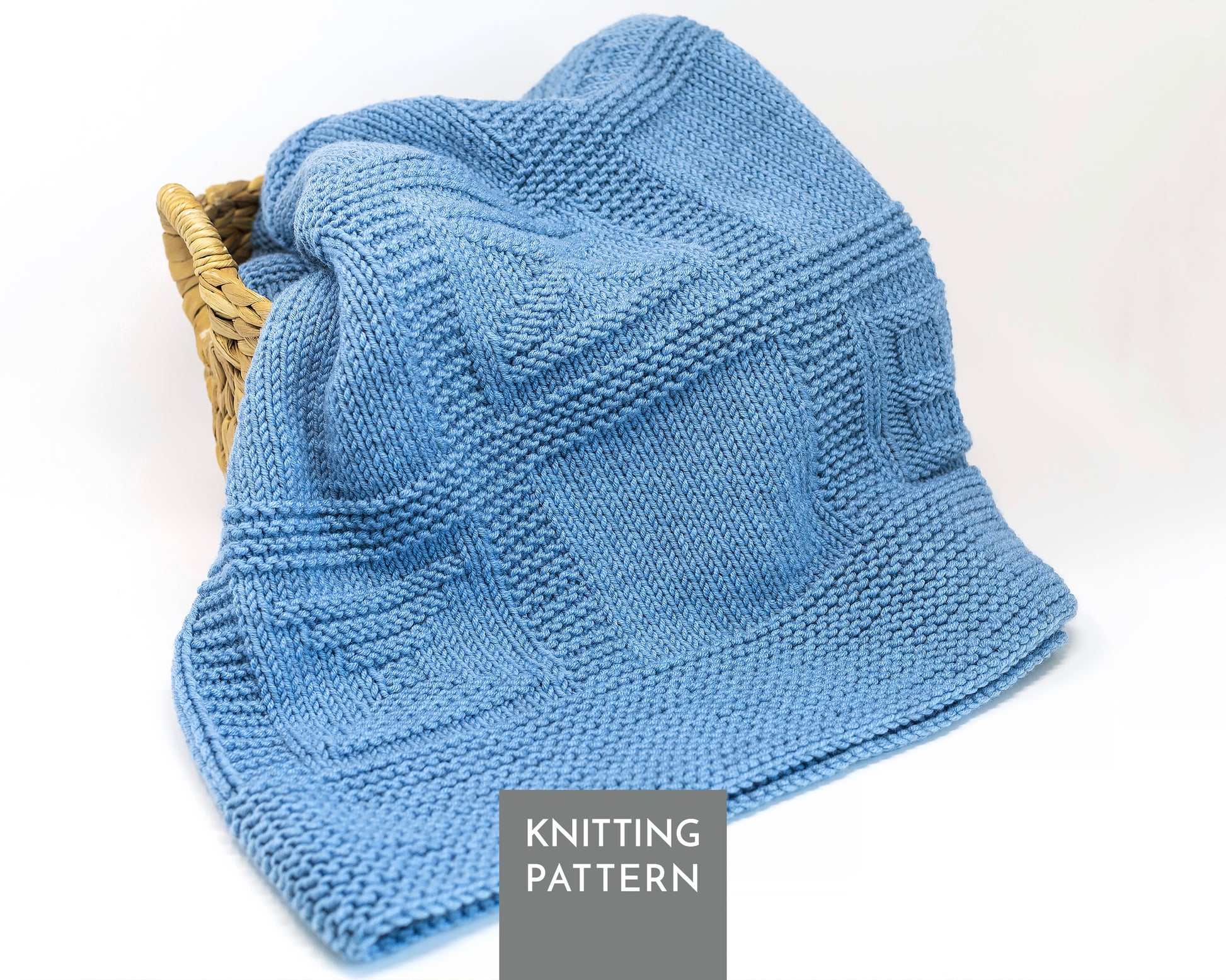 A light blue knitted baby blanket draped over a small woven basket, with a gray label reading 'KNITTING PATTERN' at the bottom. The product is shown on a clean white background.