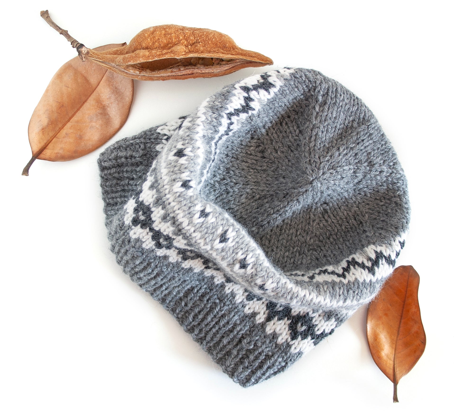 Gray and white hand-knitted Fair Isle beanie hat on a white background accompanied with dryed leaves