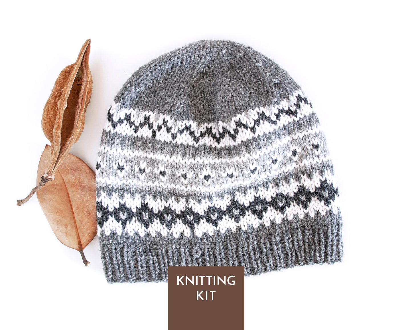 A knitted beanie hat shown on a white background next to a dried leaf, with a brown label at the bottom reading 'KNITTING KIT'. The hat has a gray top, white and gray patterned middle, and a ribbed gray bottom.