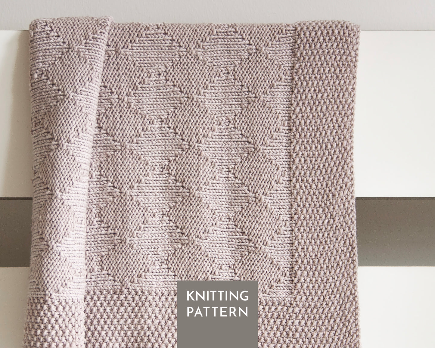 A close-up of a knitted blanket with a textured geometric diamond pattern in a beige color, draped over a white surface. A gray label at the bottom reads 'KNITTING PATTERN'.