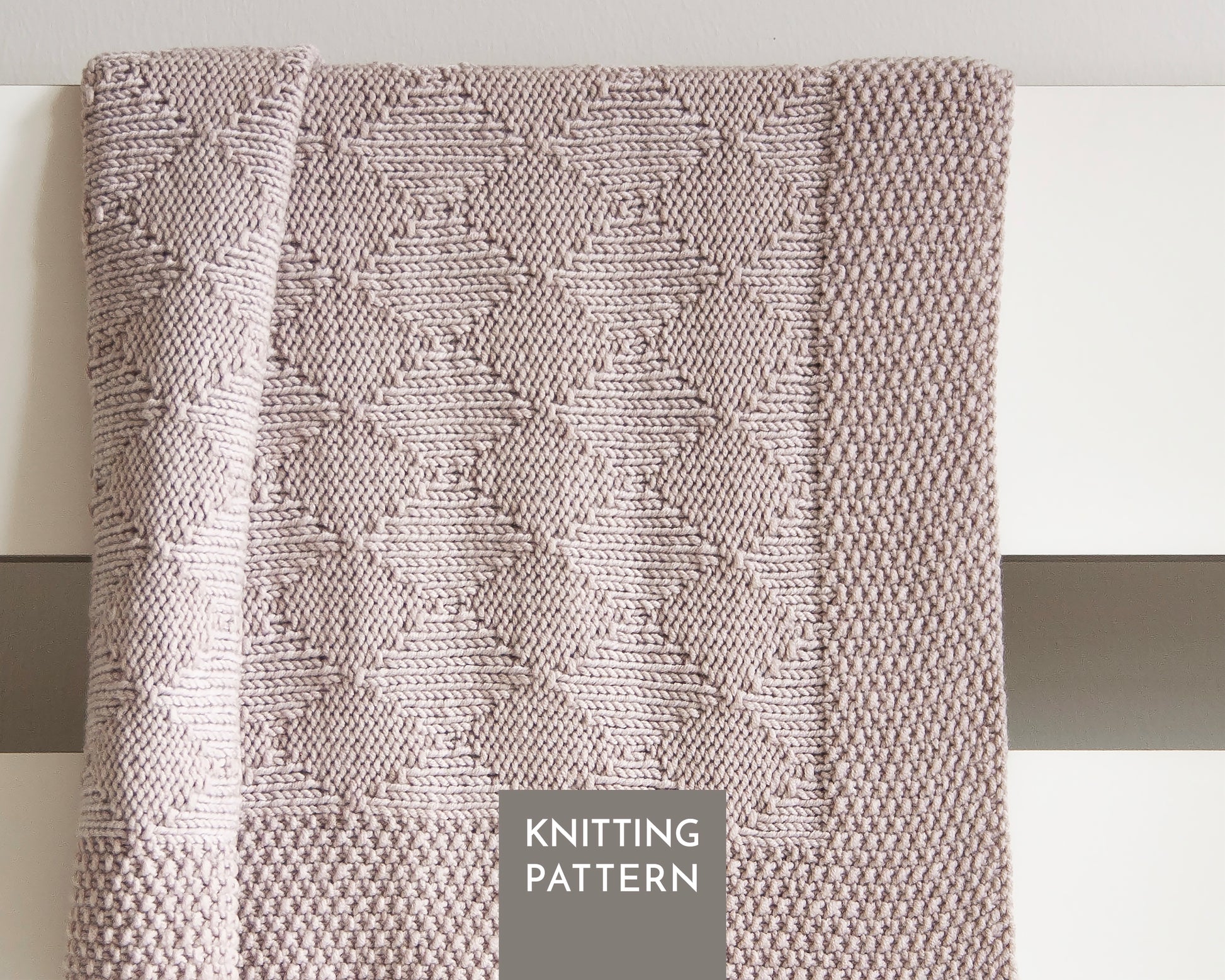 A close-up of a knitted blanket with a textured geometric diamond pattern in a beige color, draped over a white surface. A gray label at the bottom reads 'KNITTING PATTERN'.