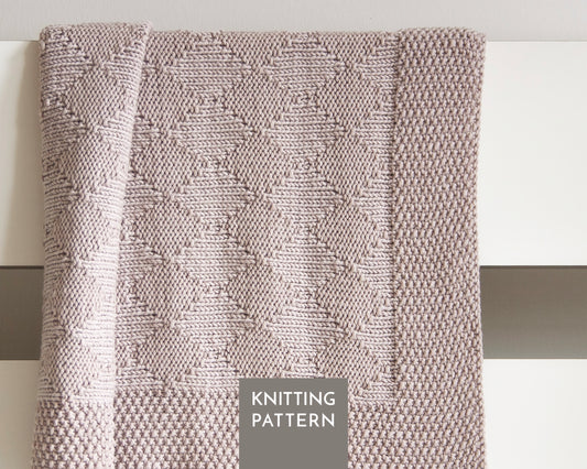 A close-up of a knitted blanket with a textured geometric diamond pattern in a beige color, draped over a white surface. A gray label at the bottom reads 'KNITTING PATTERN'.