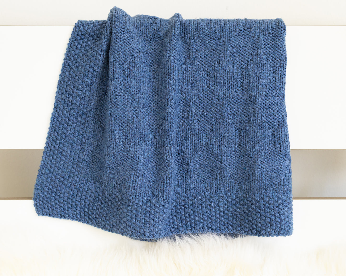 A folded blue knit blanket with a textured diamonds pattern, displayed on a white surface.