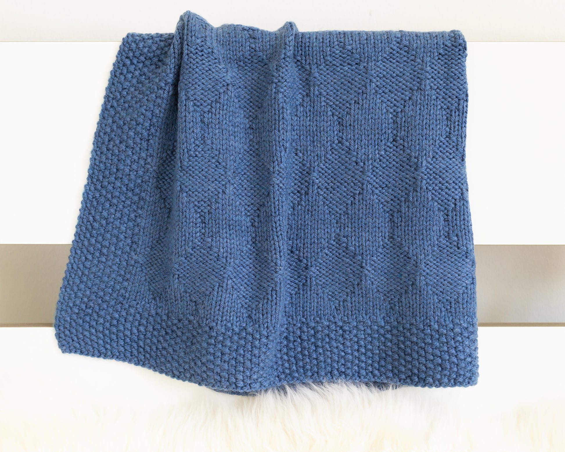 A folded blue knit blanket with a textured diamonds pattern, displayed on a white surface.