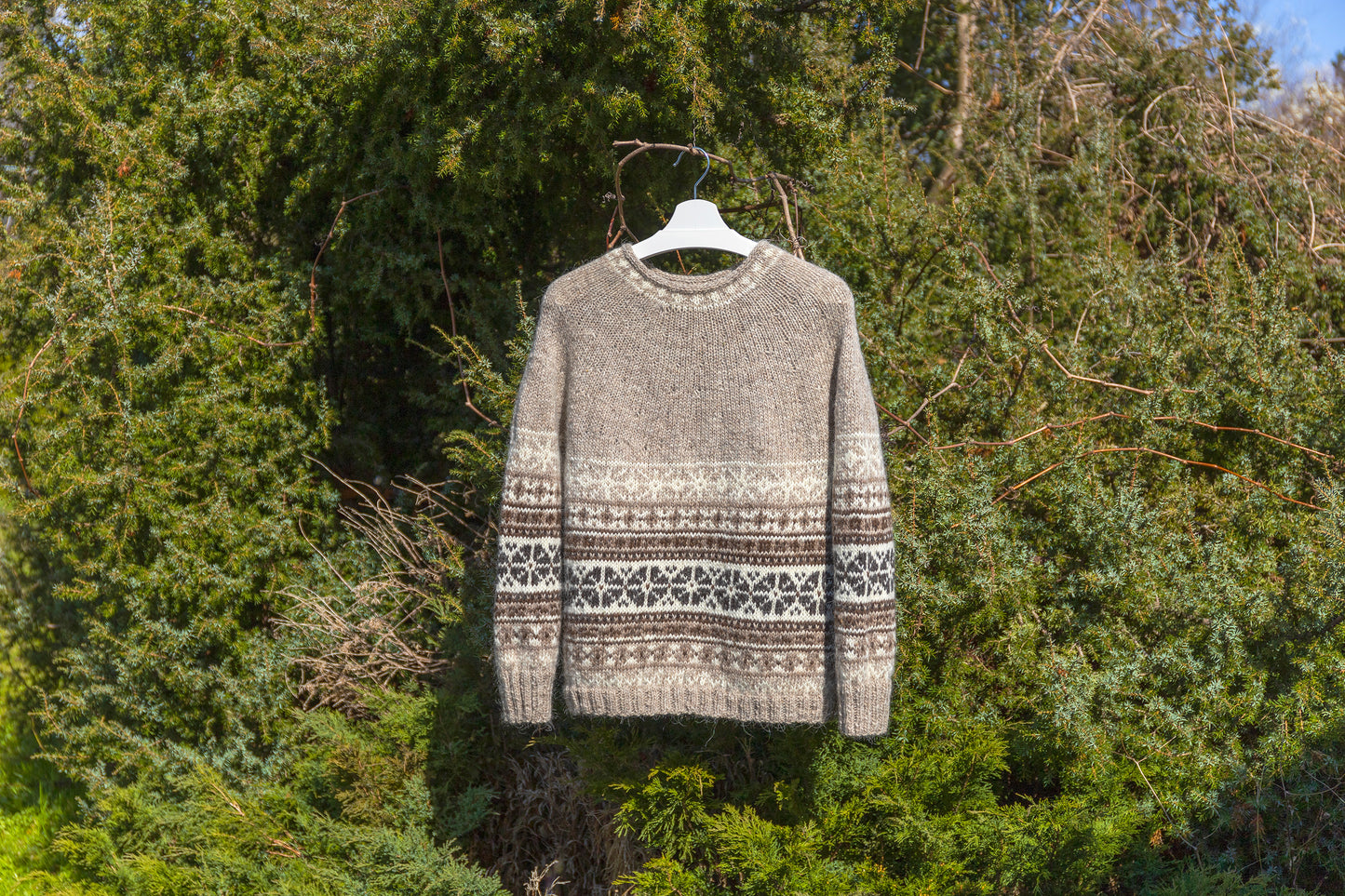 A beige and white patterned knit sweater hanging on a white hanger against a background of green trees and bushes.