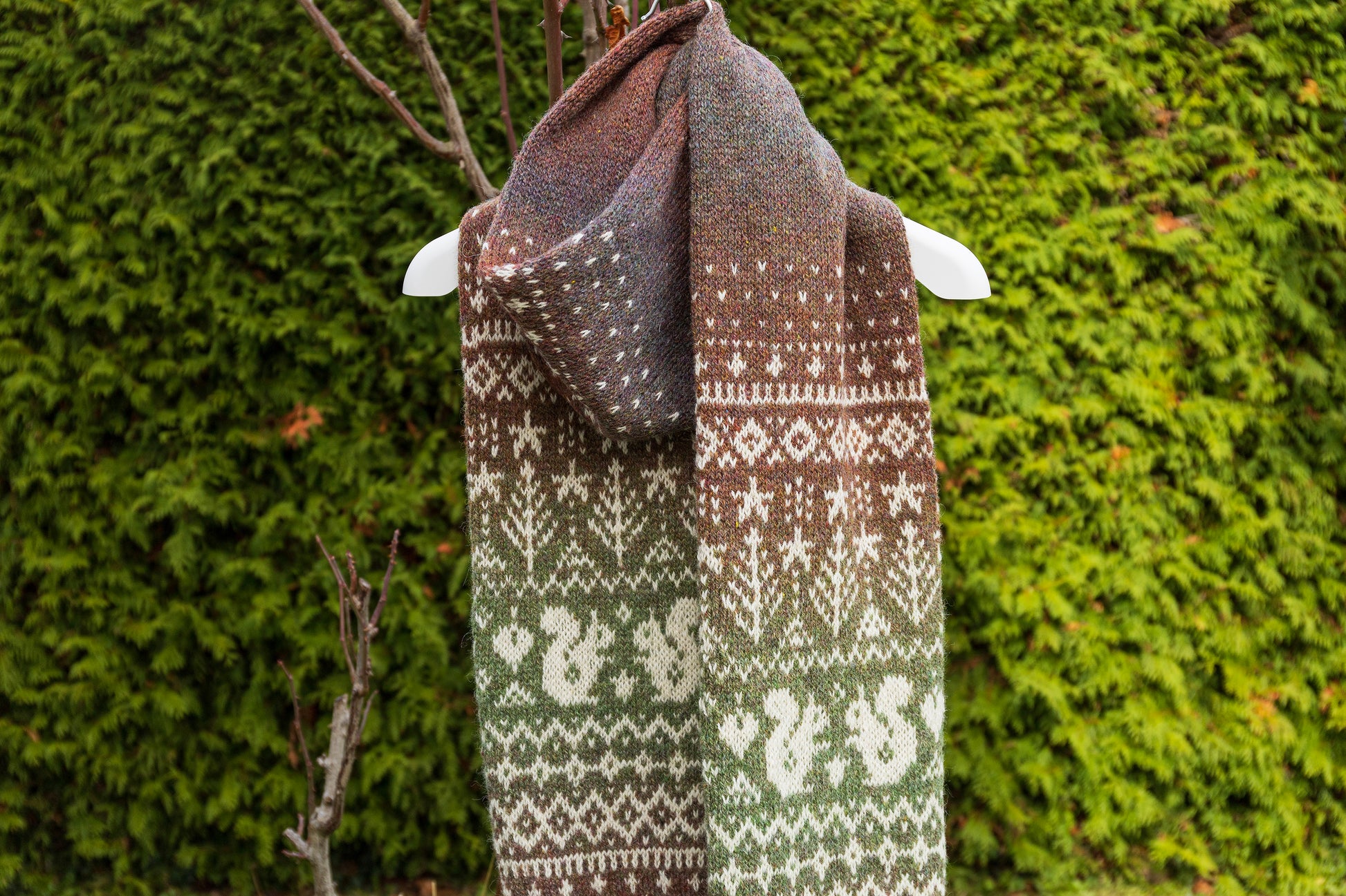 Hand-knitted Lahemaa Squirrel Fair Isle scarf draped over a branch against a green bush background