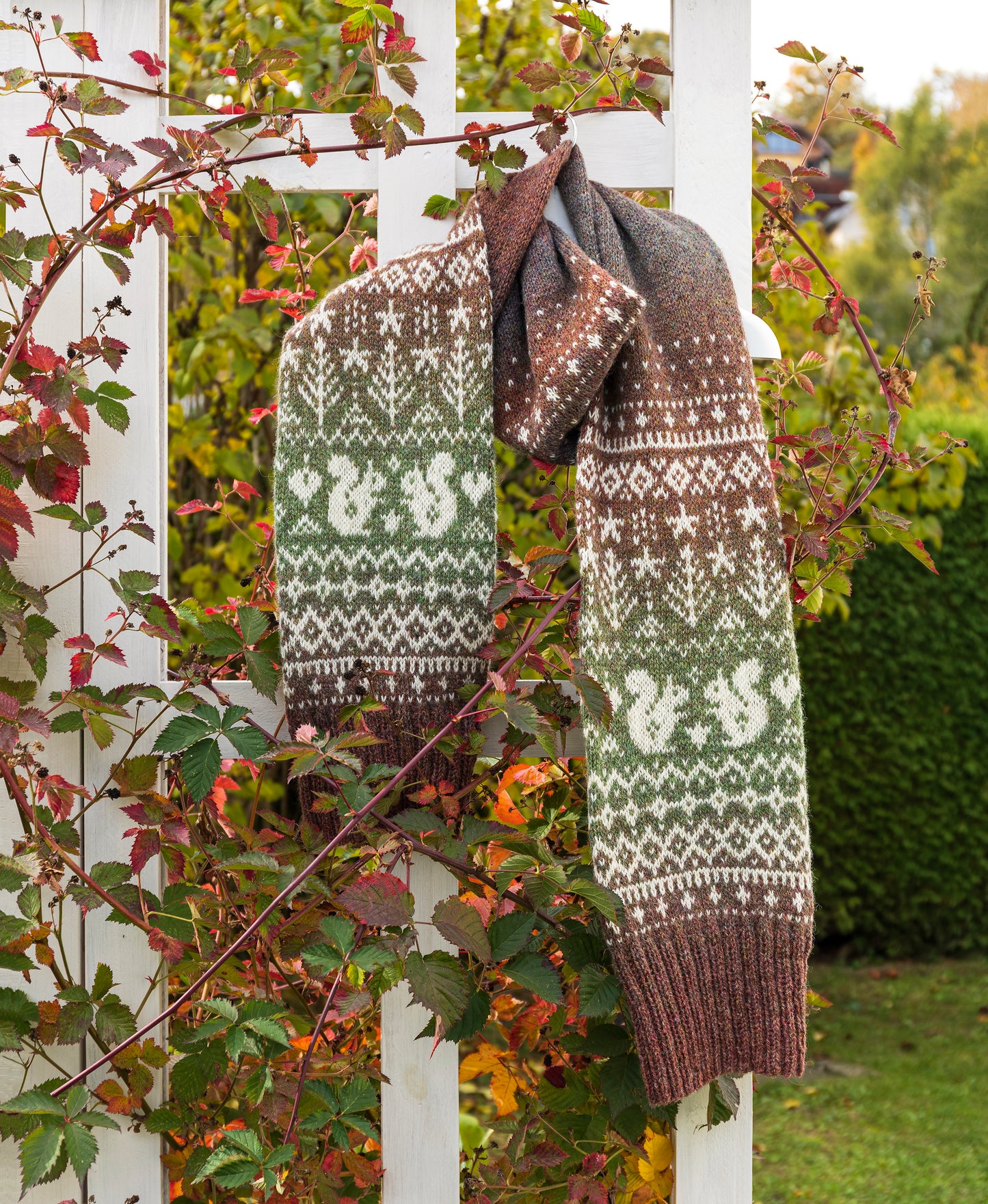 Knitted Fair Isle scarf with a squirrels and trees pattern hanging on a white fence with greenery