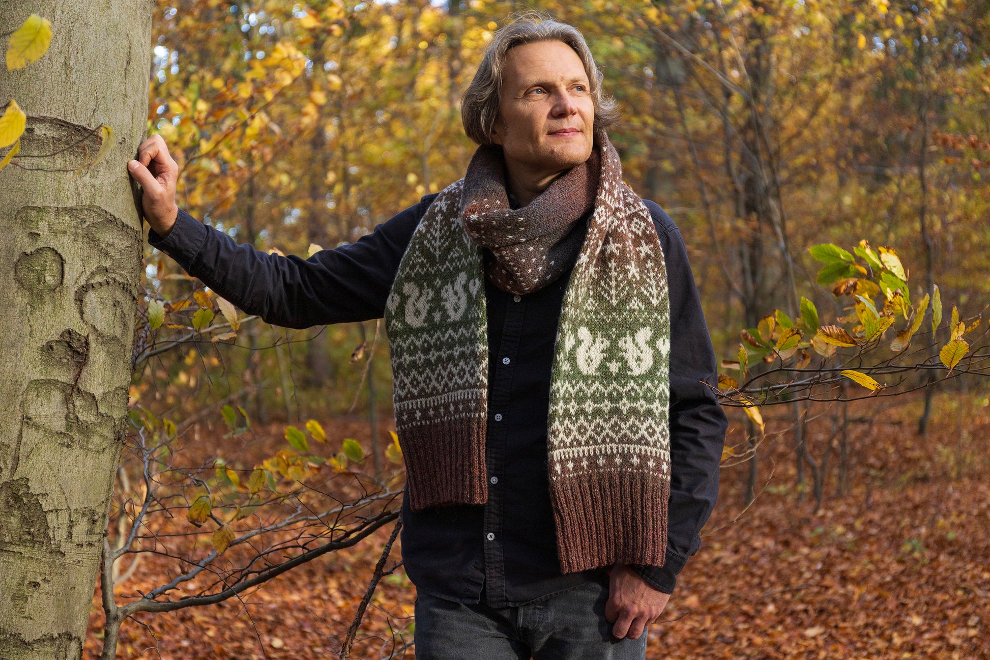 Man wearing a patterned Fair Isle scarf features squirrels, trees and stars standing in a forest with autumn leaves.