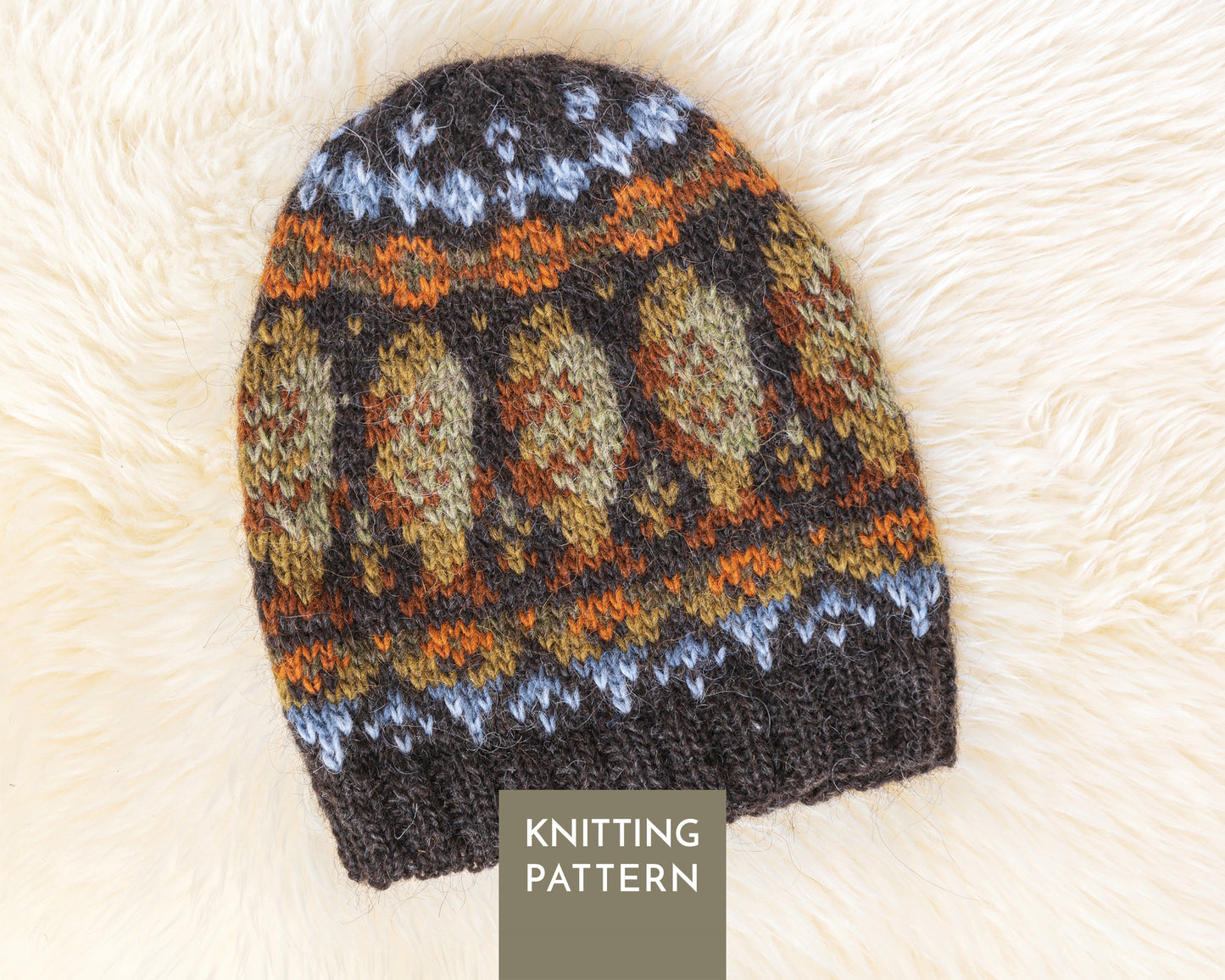 A knitted beanie hat with a Fair Isle style pattern features cod fish in dark brown, orange, beige, green and light blue, laid flat on a white fur background