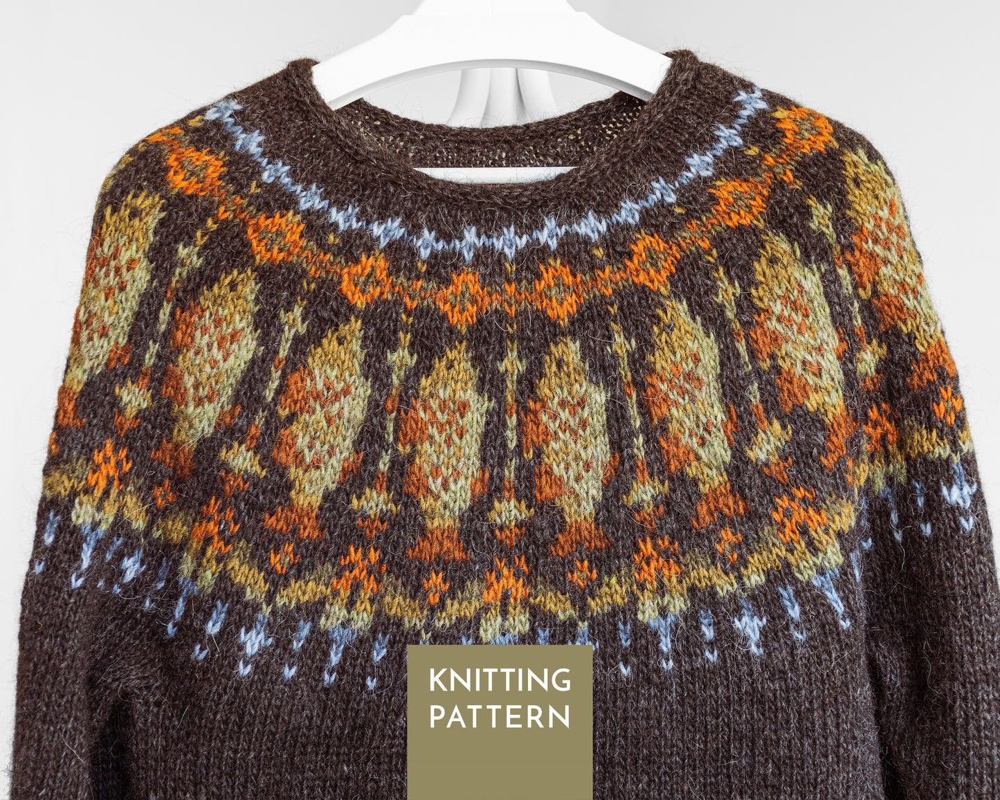 A close-up of a knitted sweater with a cod fish pattern in brown, orange, beige, and light blue, hanging on a white hanger against a light gray background.