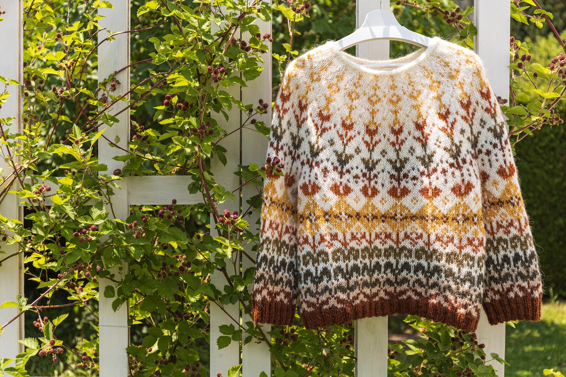 hand-knitted meadow icelandic lopapeysa sweater with floral colorwork hanging on a white railing surrounded by greenery