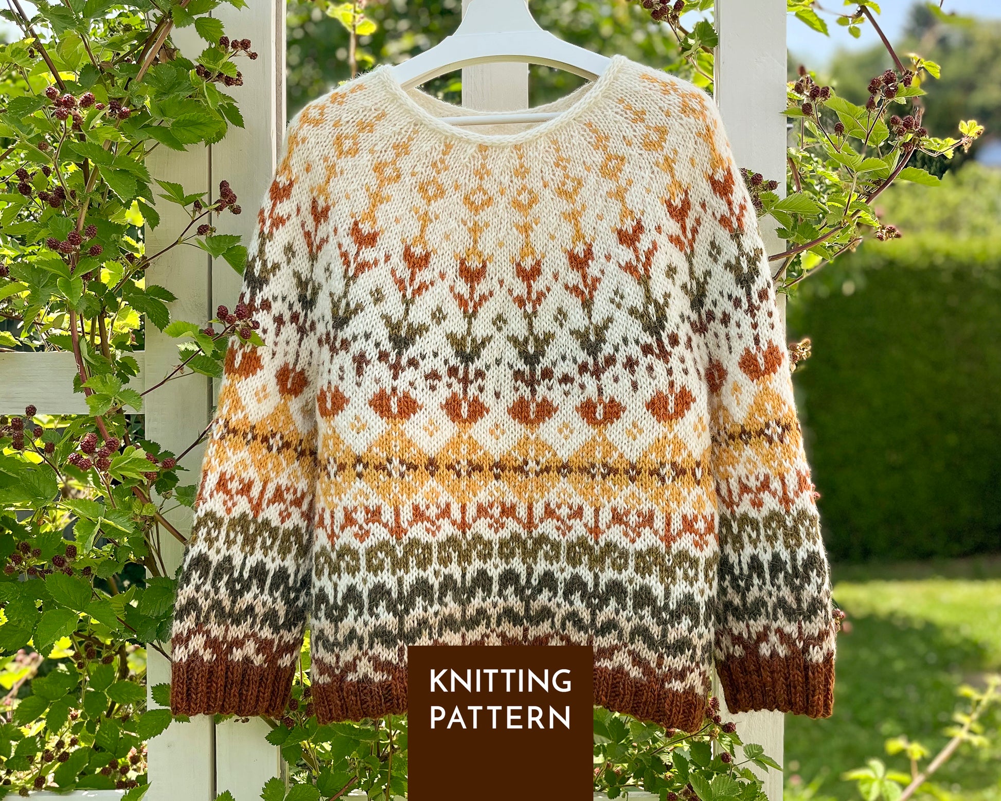 A long-sleeve knit sweater with a Fair Isle style pattern in cream, mustard yellow, rust orange, olive green, and brown, hanging on a white hanger outdoors against a white wooden fence with green foliage.
