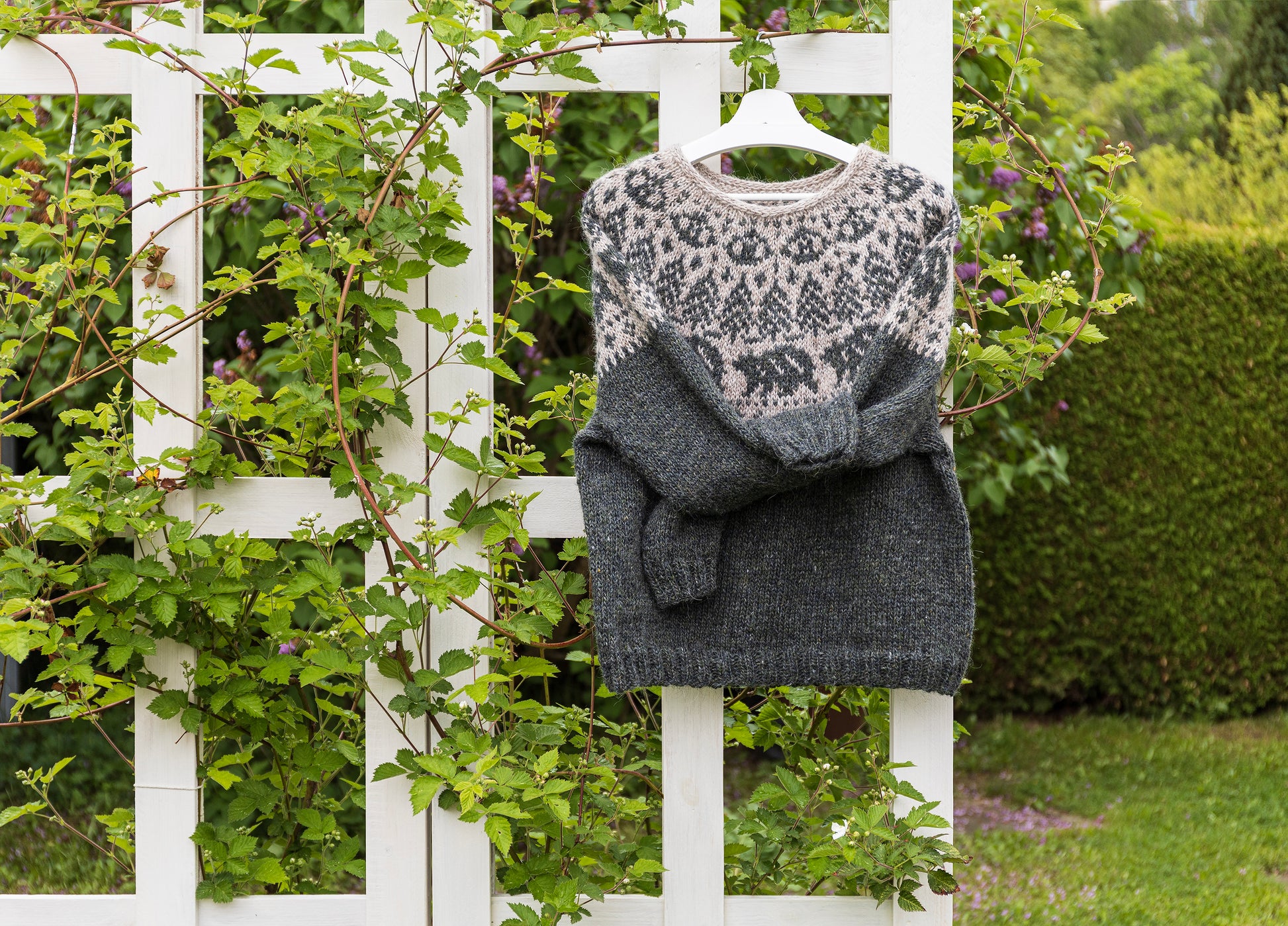 Mishka The Bear icelandic lopapeysa sweater on a hanger against a white picket fence with greenery.