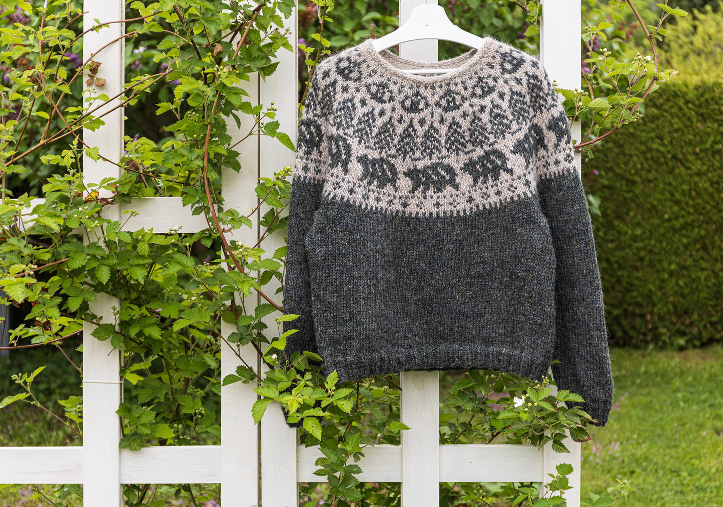 A hand-knitted Icelandic Lopapeysa sweater featuring a bear motif, displayed on a white lattice against a backdrop of greenery.