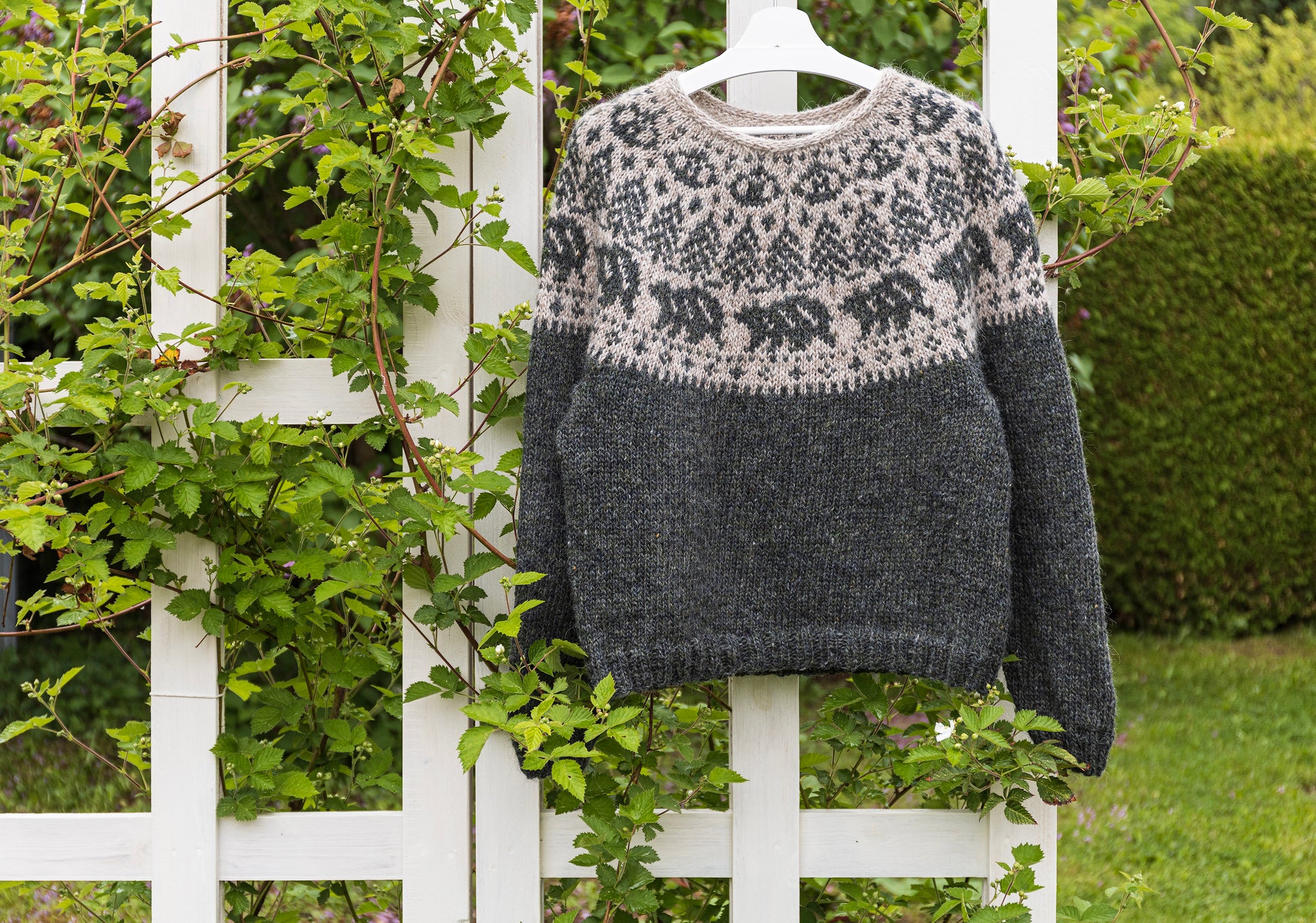 A hand-knitted Icelandic Lopapeysa sweater featuring a bear motif, displayed on a white lattice against a backdrop of greenery.