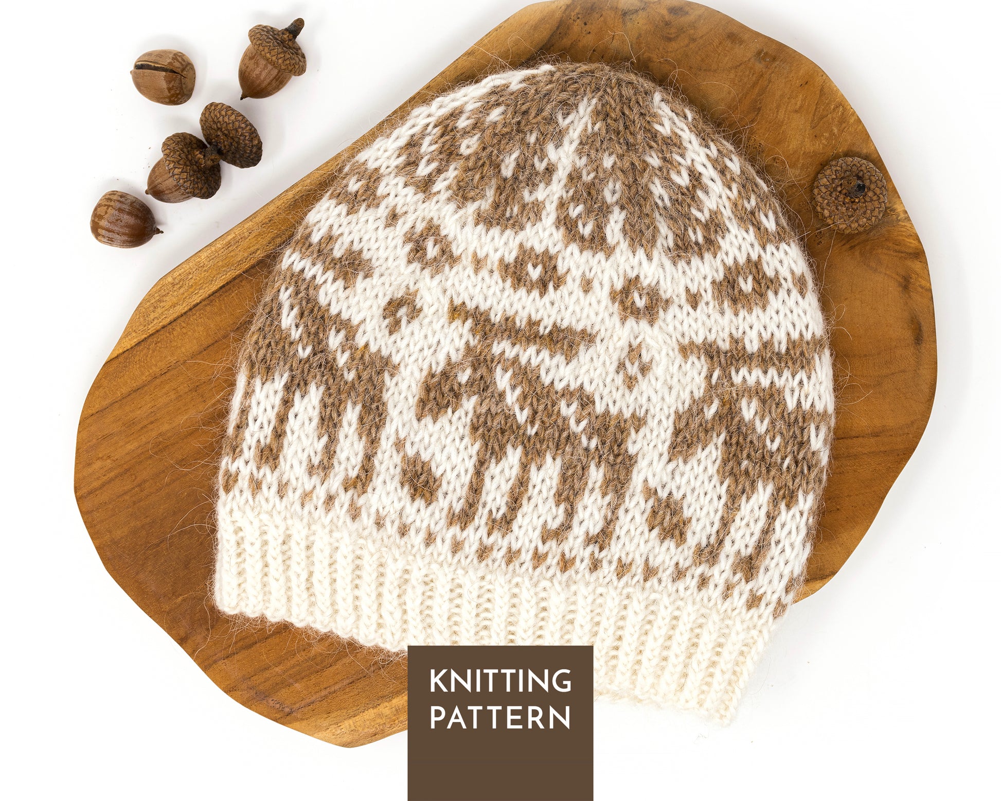 A knitted beanie hat with a brown and cream fair isle pattern features moose, displayed on a rustic wooden board with acorns