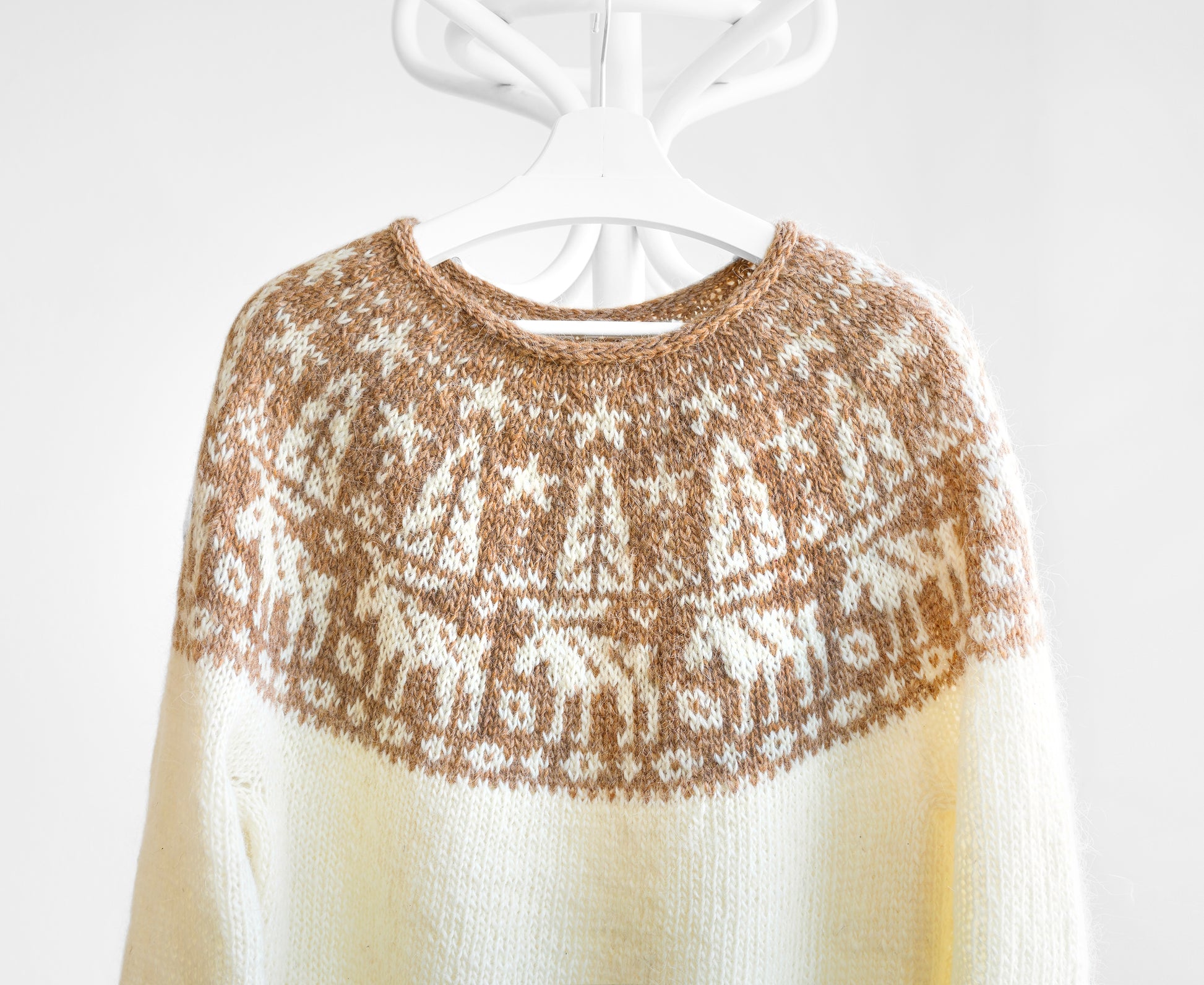 A cream-coloured lopapeysa sweater featuring a round yoke with a brown and white fair isle pattern depicting stylised mooses, trees, and stars.