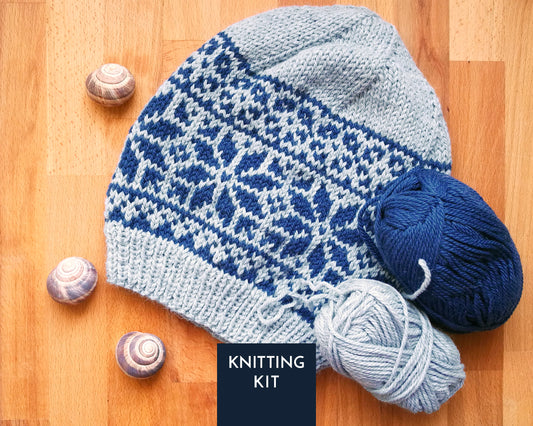 A knitted beanie hat with a blue and light gray Fair Isle pattern, displayed on a wooden surface alongside balls of yarn and small seashells, with a 'KNITTING KIT' label overlay.